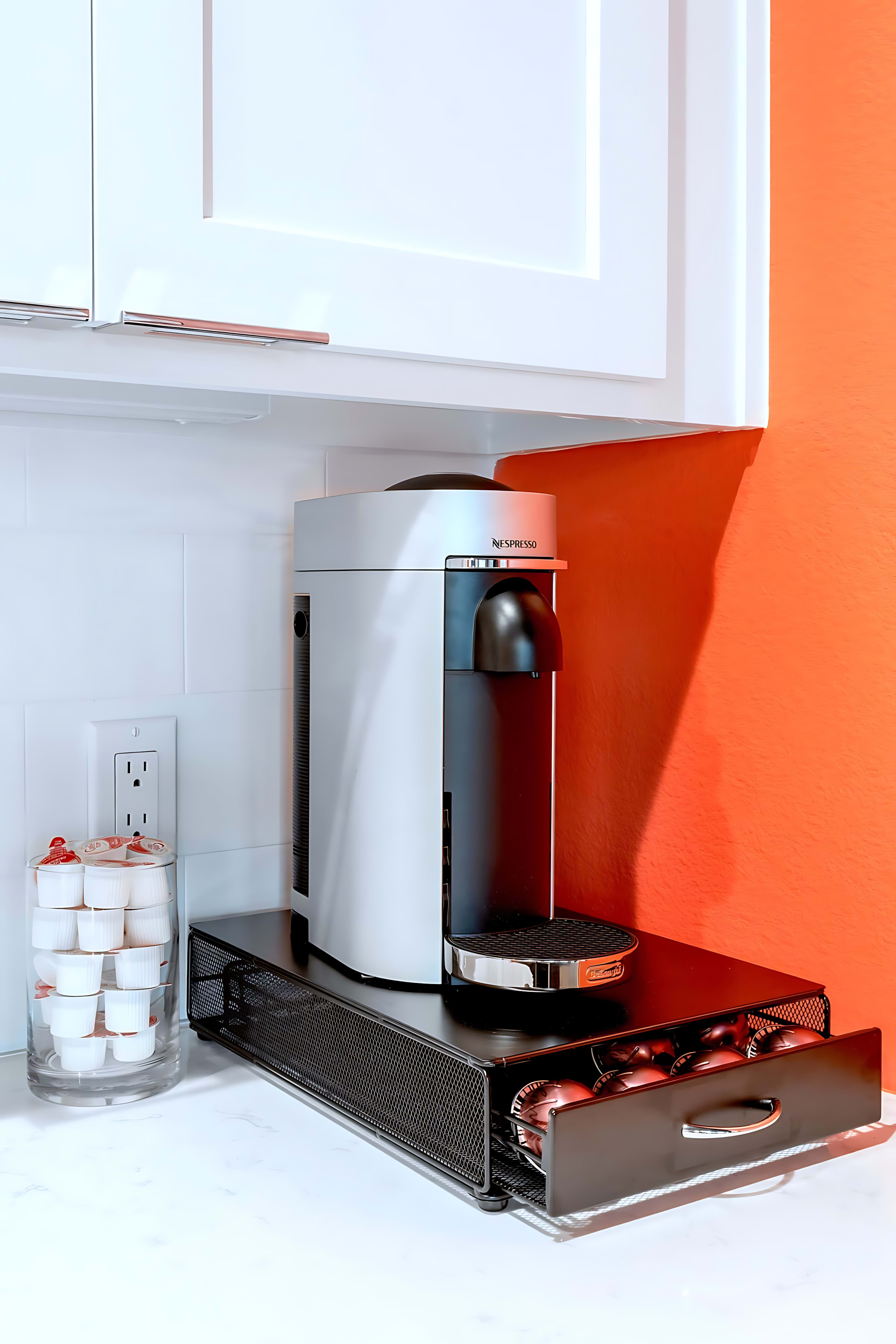 A Nespresso coffee machine sits on a white countertop next to a clear jar of coffee pods, with a black mesh drawer organizer for more pods underneath, against an orange wall.