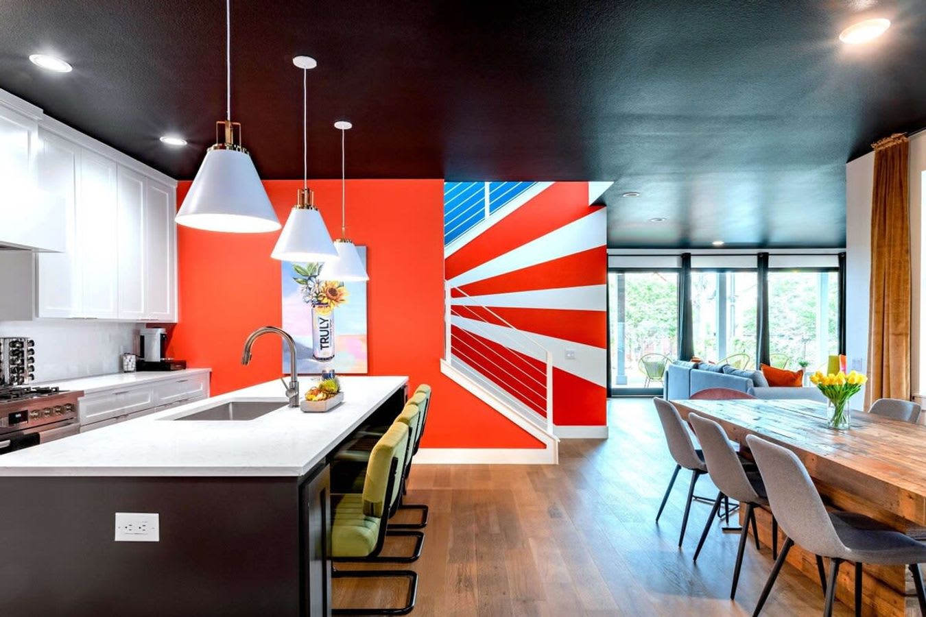 Open-concept kitchen and dining area with a large kitchen island, white cabinets, and an orange accent wall, leading to a dining space and a striking red, white, and blue striped staircase.
