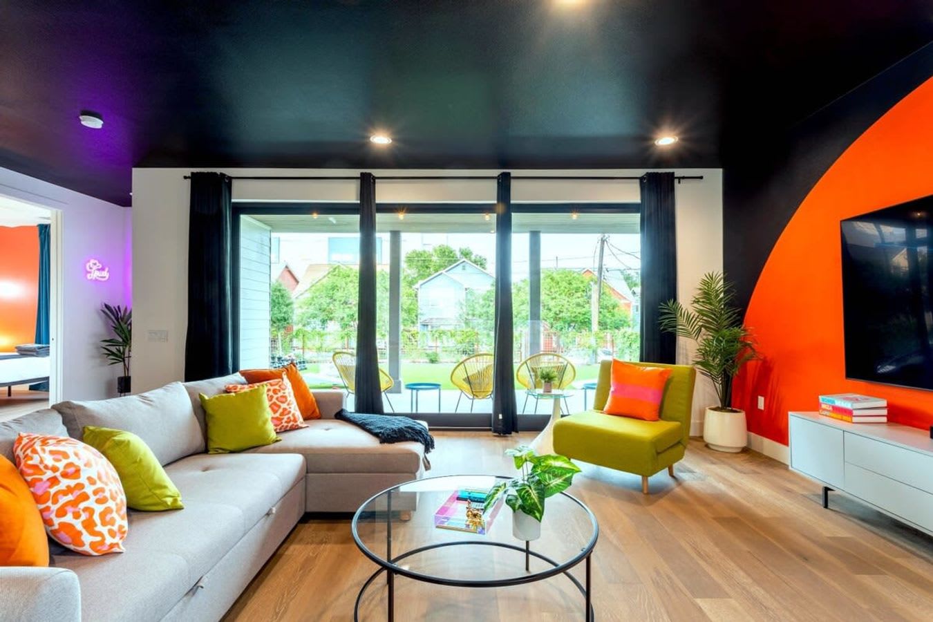 Vibrant living room with a black ceiling, orange accent wall, and a large grey sectional sofa with colorful pillows, facing a glass coffee table and large sliding glass doors leading to an outdoor patio.