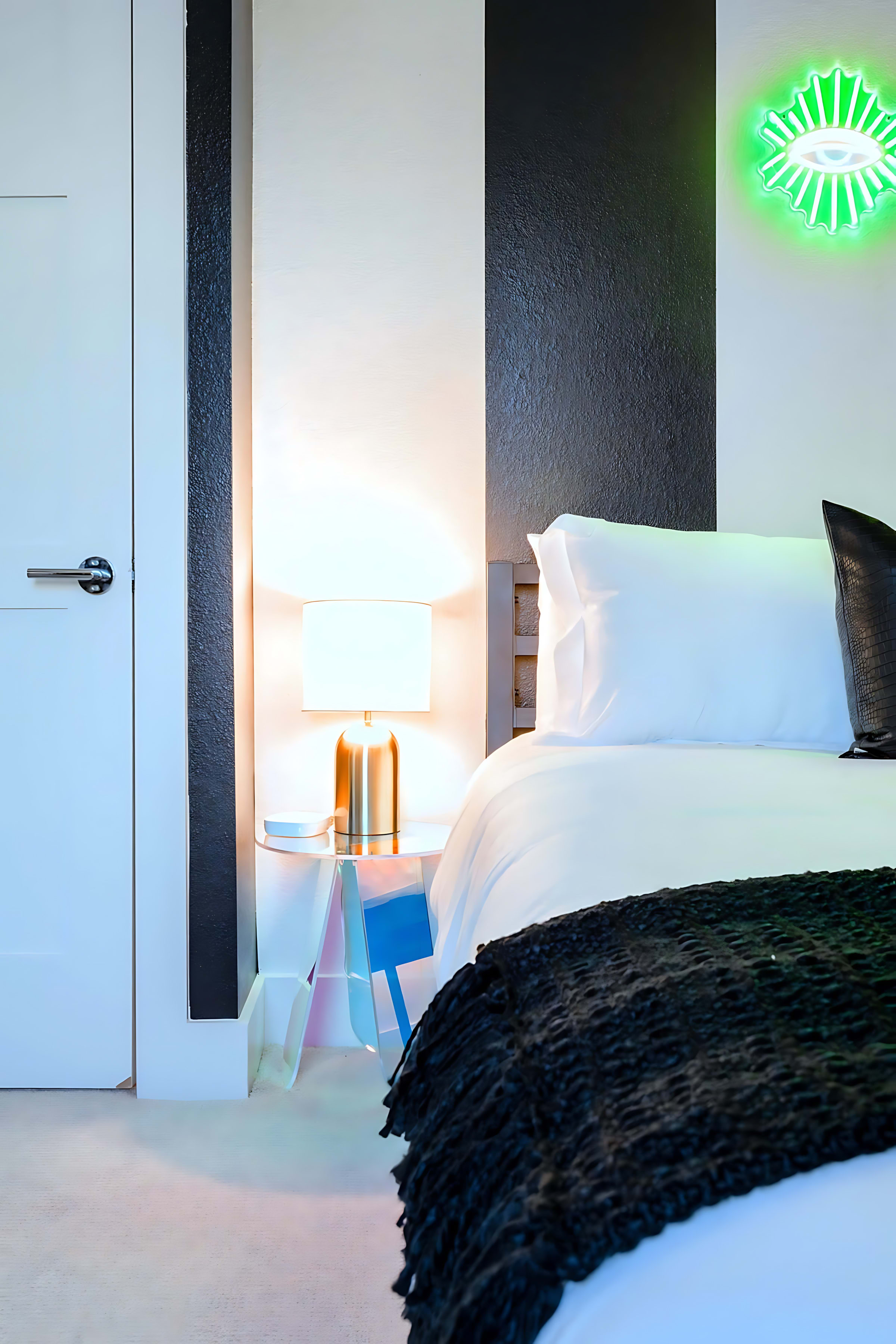 A close-up of a modern bedroom showing a white bed with a black throw and a black textured pillow, next to a clear acrylic nightstand with a gold-based lamp. The wall features bold black and white vertical stripes, and a green neon eye art piece is visible.