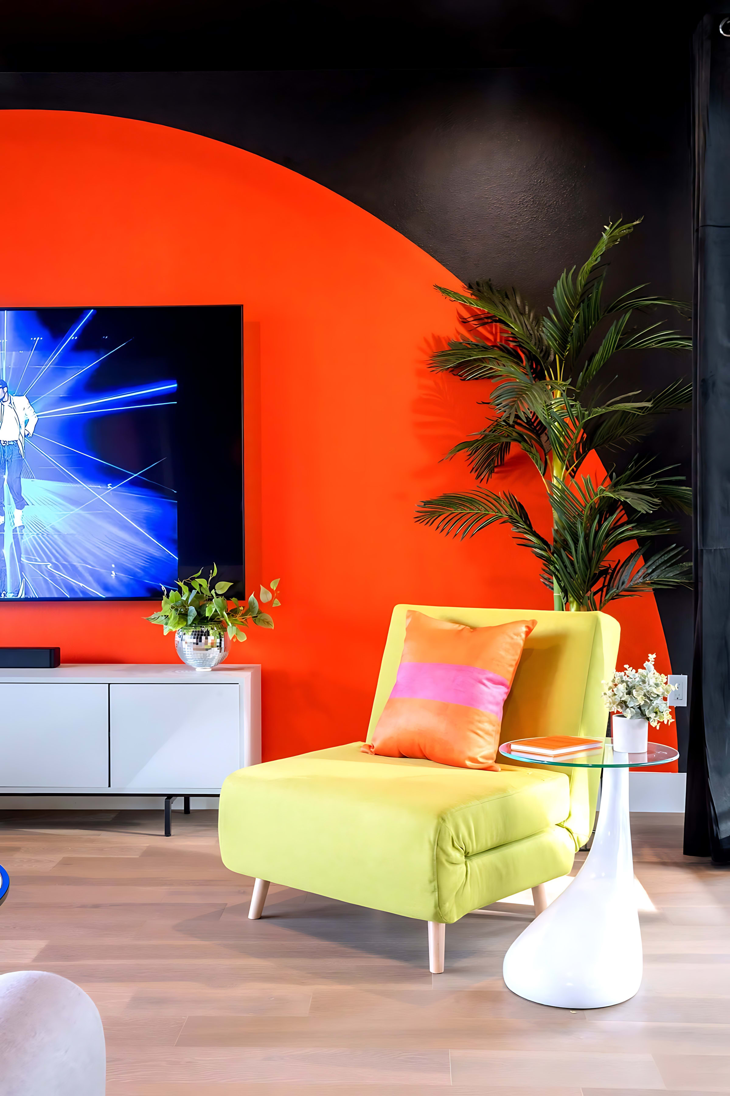A bright lime green armchair with an orange and pink pillow sits beside a glass side table, against a bold orange accent wall with a wall-mounted TV and a tall potted plant.