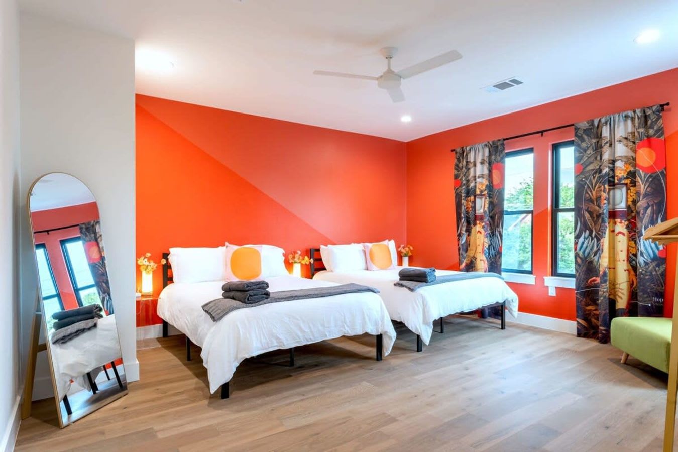 A vibrant bedroom with two white-linened beds, each featuring an orange circular pillow, set against a striking orange accent wall with a diagonal lighter orange stripe. A full-length mirror stands on the left, reflecting the room, and a ceiling fan is visible above.