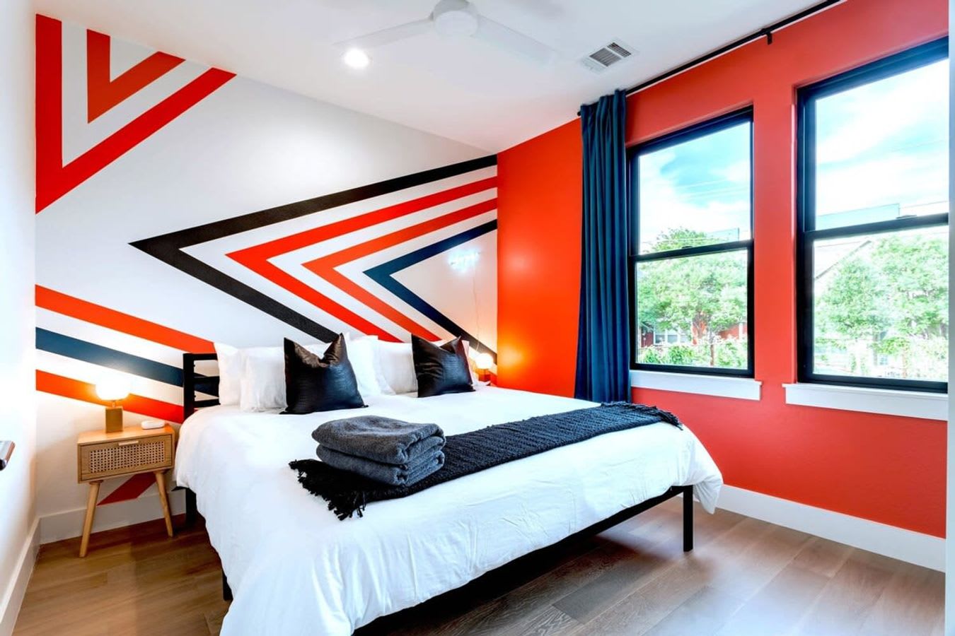 A modern bedroom featuring a white bed with black pillows and throw, set against a white wall with bold red, black, and blue geometric stripes. A large window with blue curtains offers a view of trees, and a wooden nightstand with a lamp is visible.