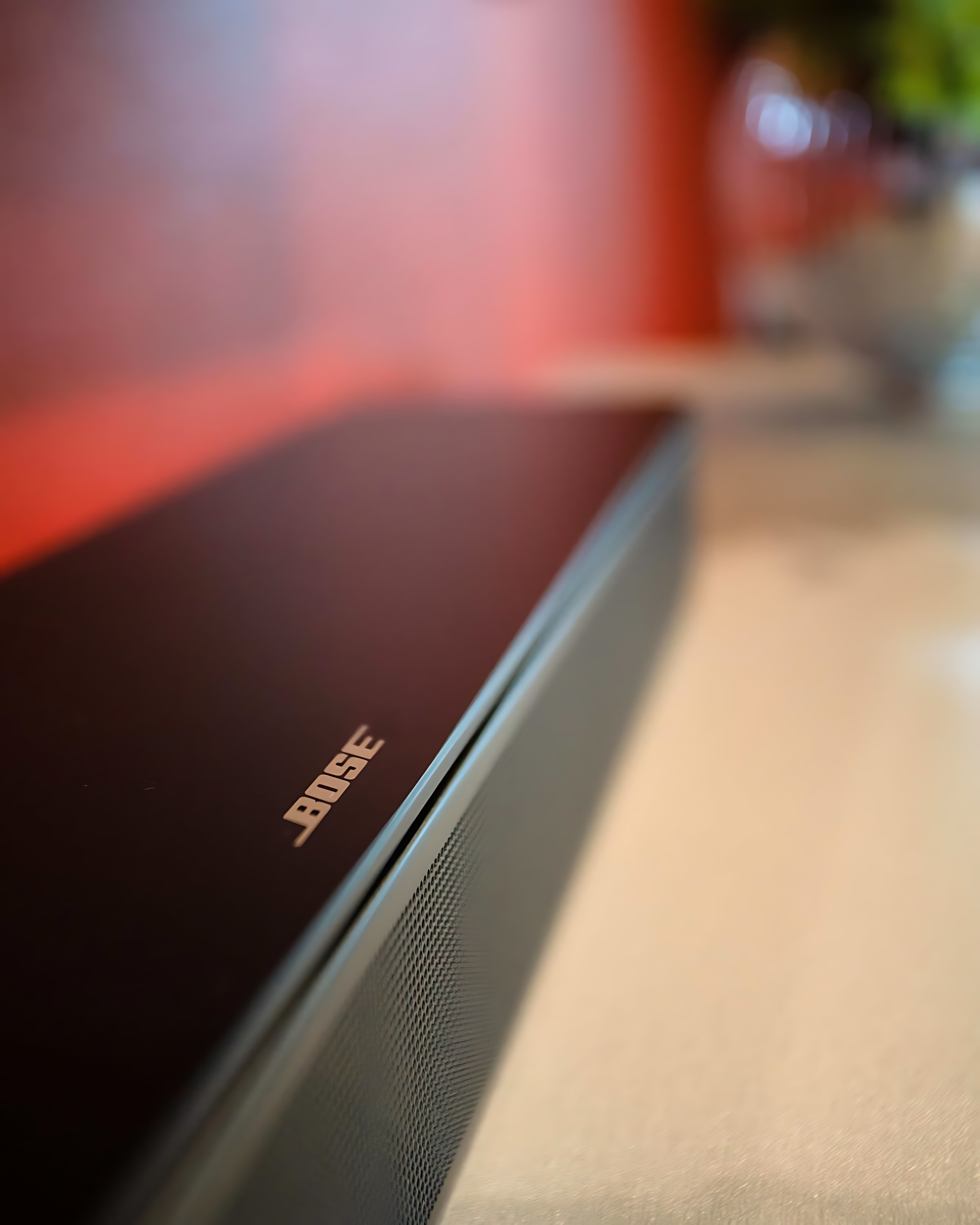 A close-up shot of a black Bose soundbar with the brand logo visible, resting on a light-colored surface, with a blurred orange wall in the background.