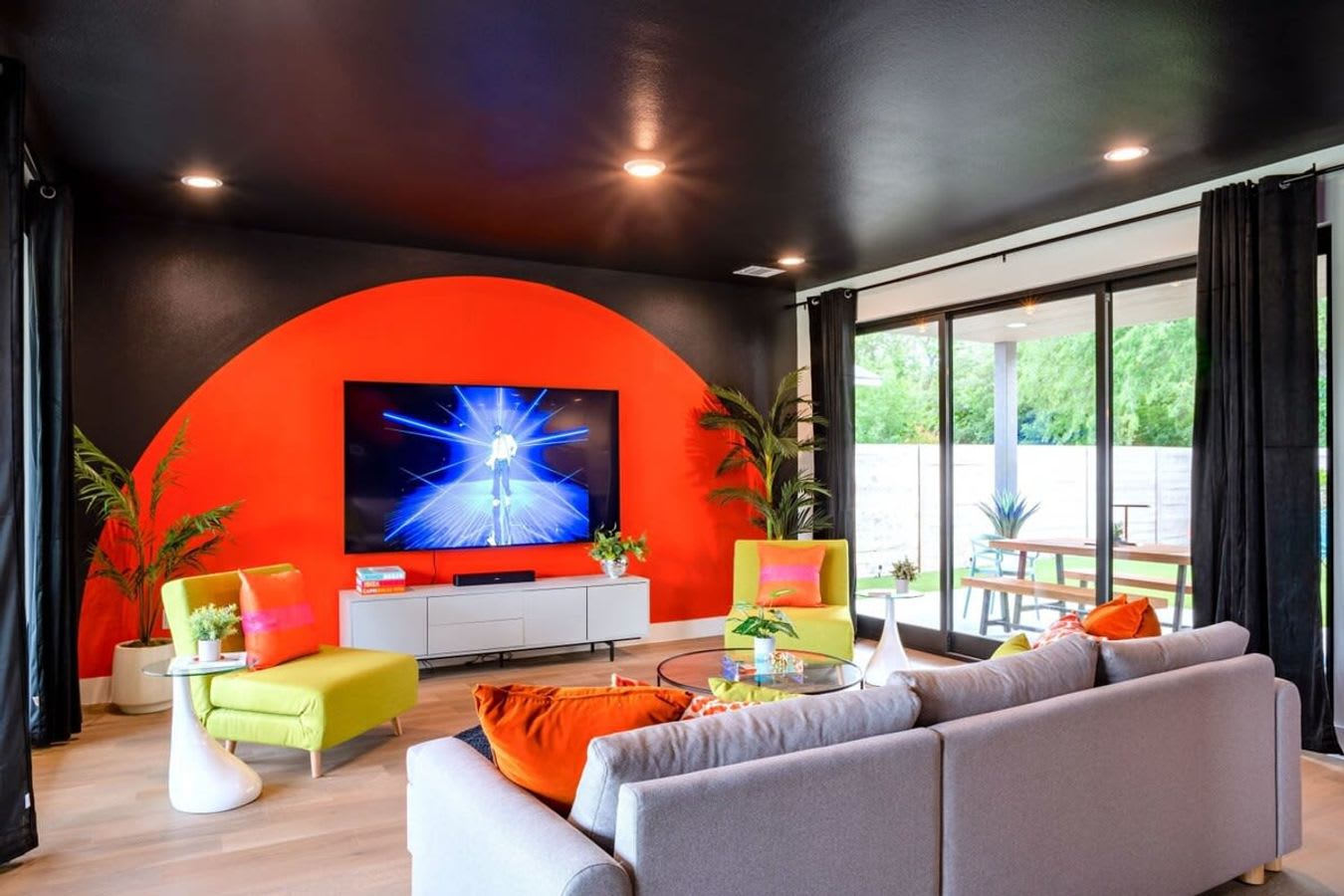 Spacious living room featuring a large grey sectional sofa, two lime green armchairs, and a wall-mounted TV on a vibrant orange accent wall, all under a black ceiling with recessed lighting.