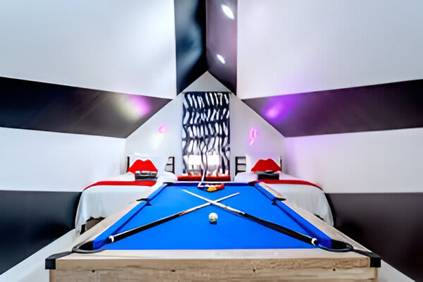 A unique attic game room features a blue-felt pool table at its center, flanked by two beds with distinctive red lip pillows, all under a striking black and white striped ceiling and walls with purple accent lighting.