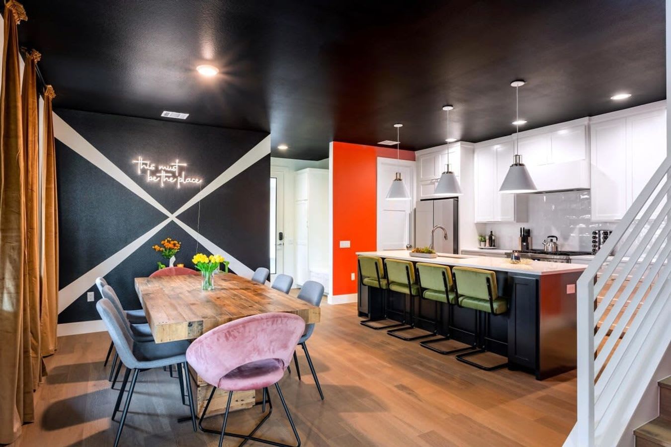 A stylish dining area with a rustic wooden table and mixed seating, including gray and pink velvet chairs, is illuminated by a neon sign on a black and white accent wall, adjacent to a modern kitchen with a large island and green bar stools.