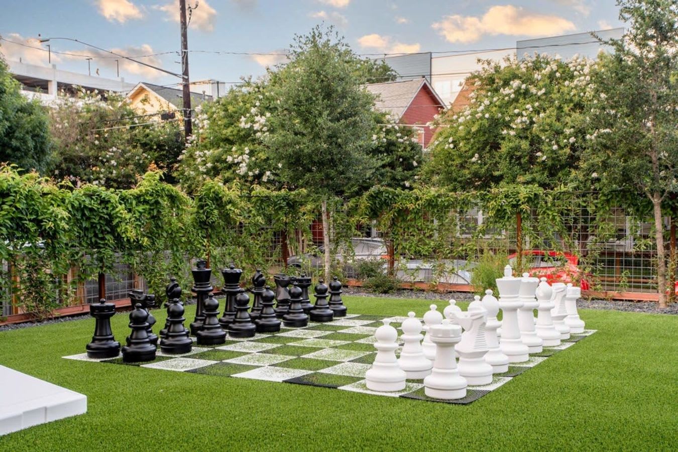 A large outdoor giant chess set with black and white pieces is laid out on a checkered green and white mat on artificial turf, surrounded by lush green foliage and a wooden fence in the background.