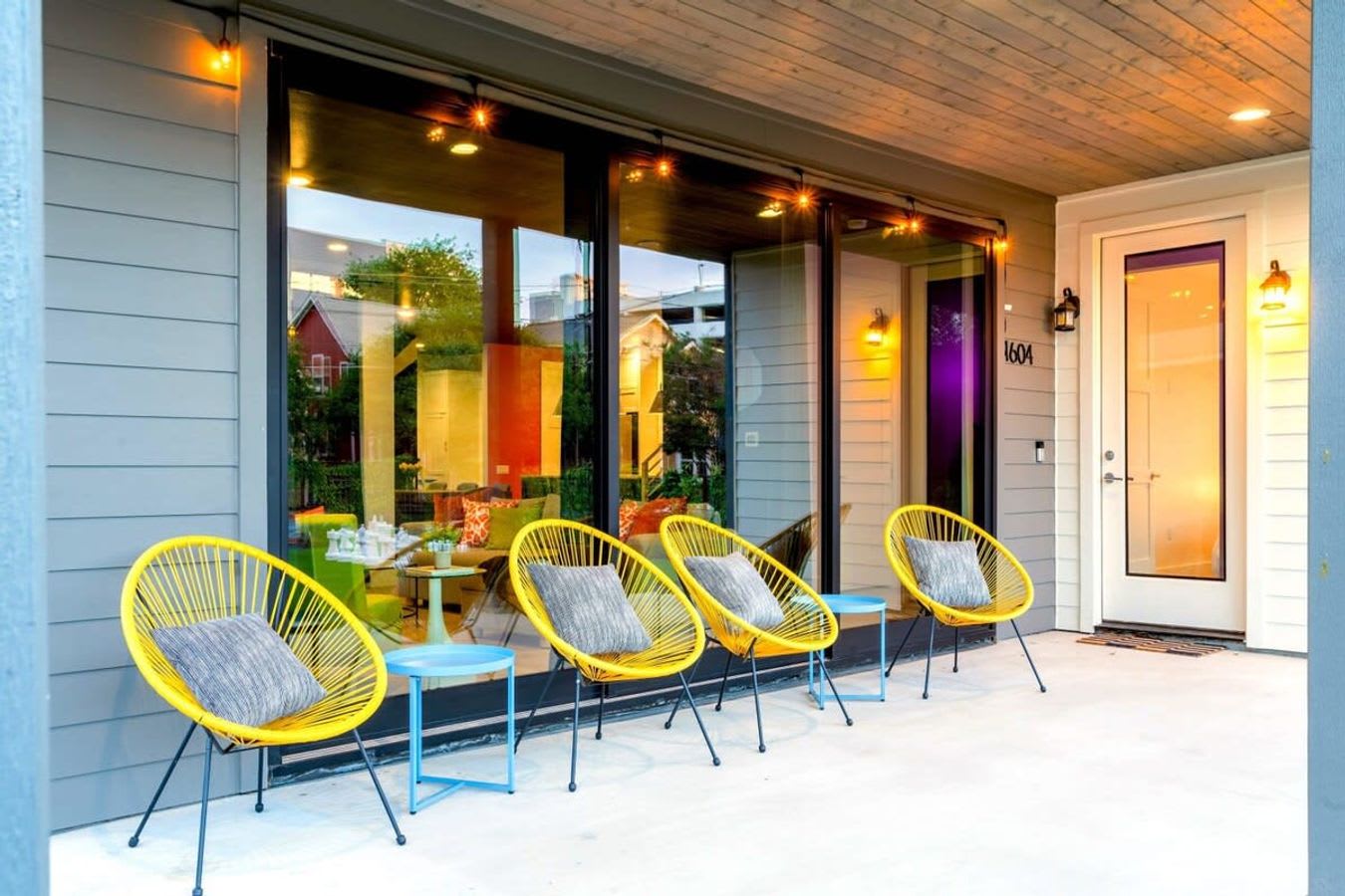 A welcoming covered porch features a row of four bright yellow string chairs with gray pillows and small blue side tables, set against a modern house exterior with large glass sliding doors and warm interior lighting.