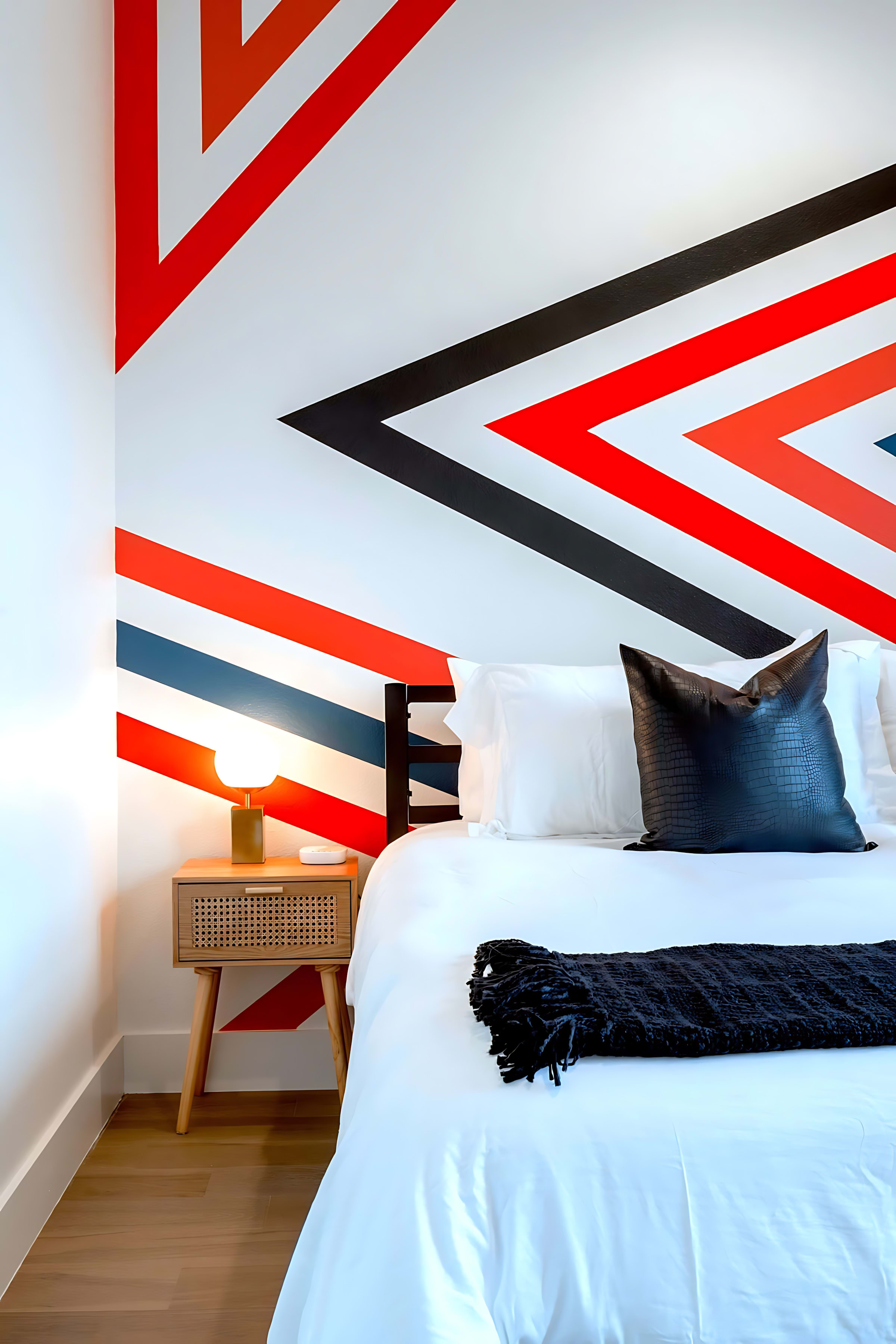 A close-up of a modern bedroom showing a white bed with a black throw and a black textured pillow, next to a wooden nightstand with a small lamp. The wall behind features a striking geometric pattern in red, black, and blue on a white background.