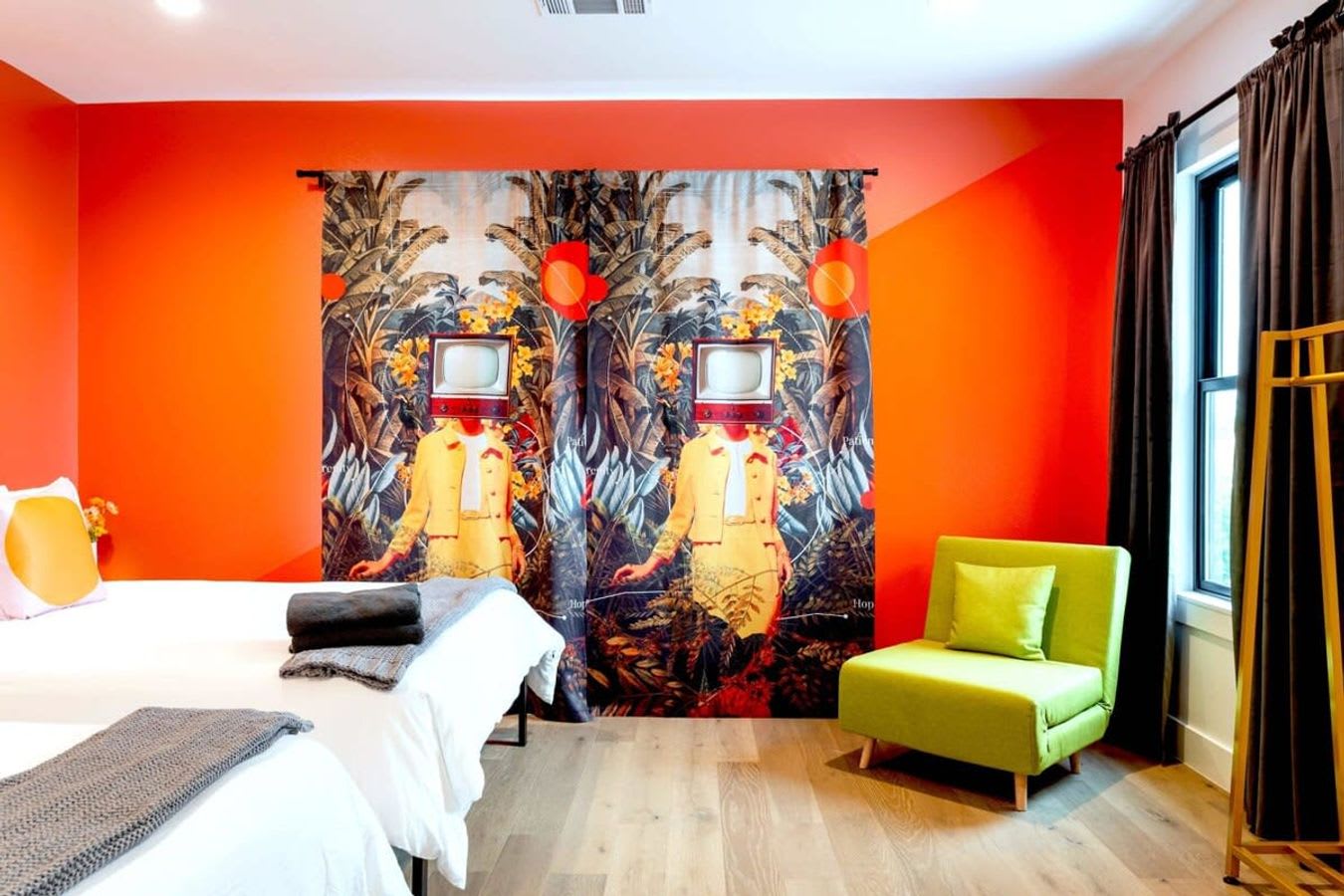 A wide shot of a vibrant bedroom featuring two white-linened beds with orange pillows, set against an orange accent wall. Large windows are adorned with unique, colorful curtains depicting figures with TV heads, and a lime green armchair sits in the corner.