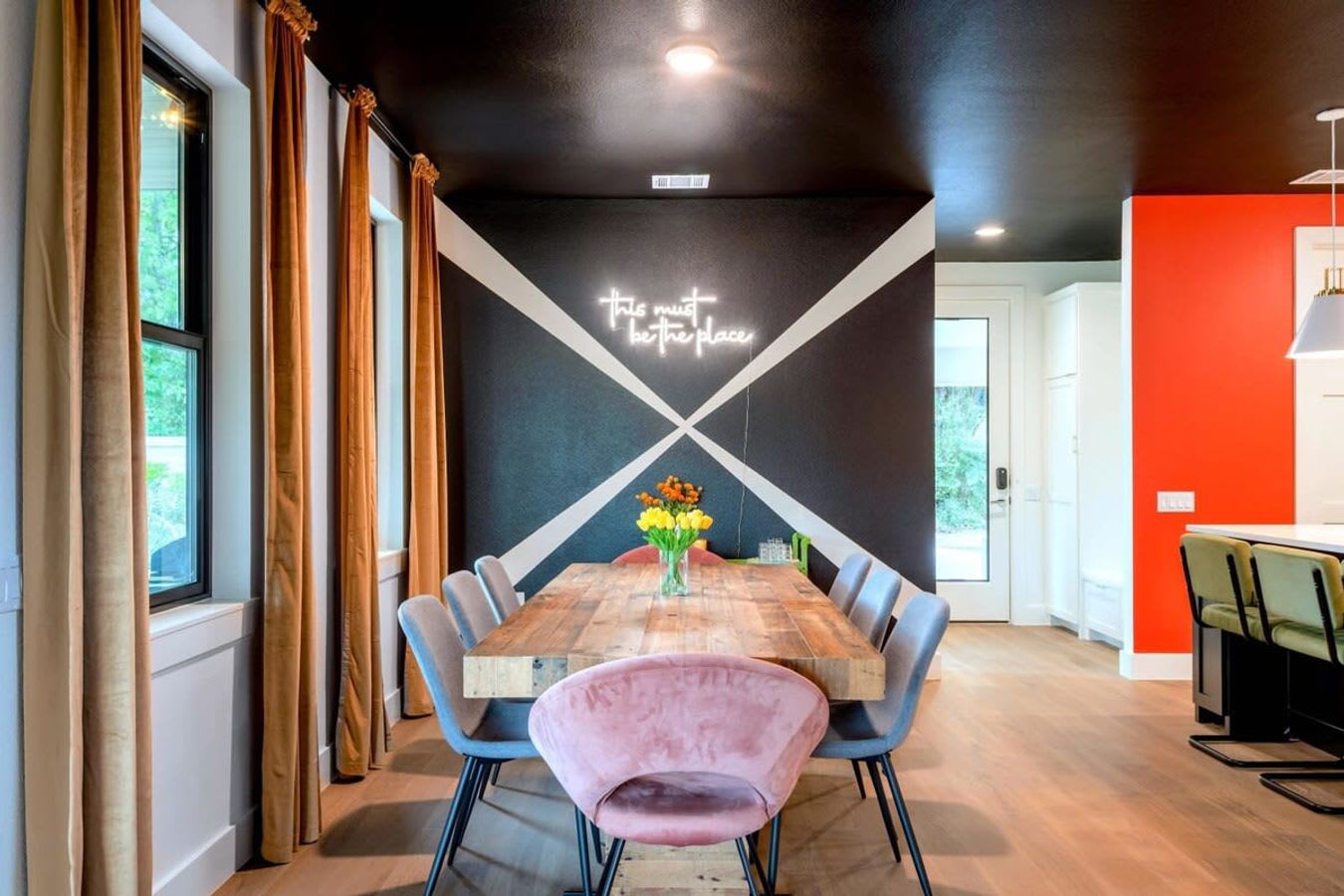 Modern dining room with a long rustic wooden table surrounded by grey and pink velvet chairs, set against a black wall with a white geometric pattern and a neon sign, under a black ceiling.
