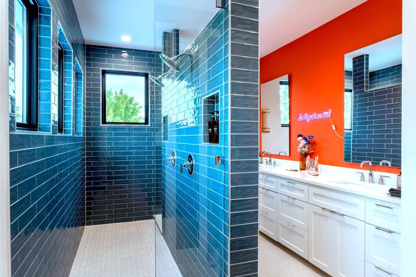 A spacious walk-in shower with glossy blue subway tiles covering the walls and a small window. The shower features multiple showerheads and a glass partition, with the double vanity and orange accent wall visible beyond.