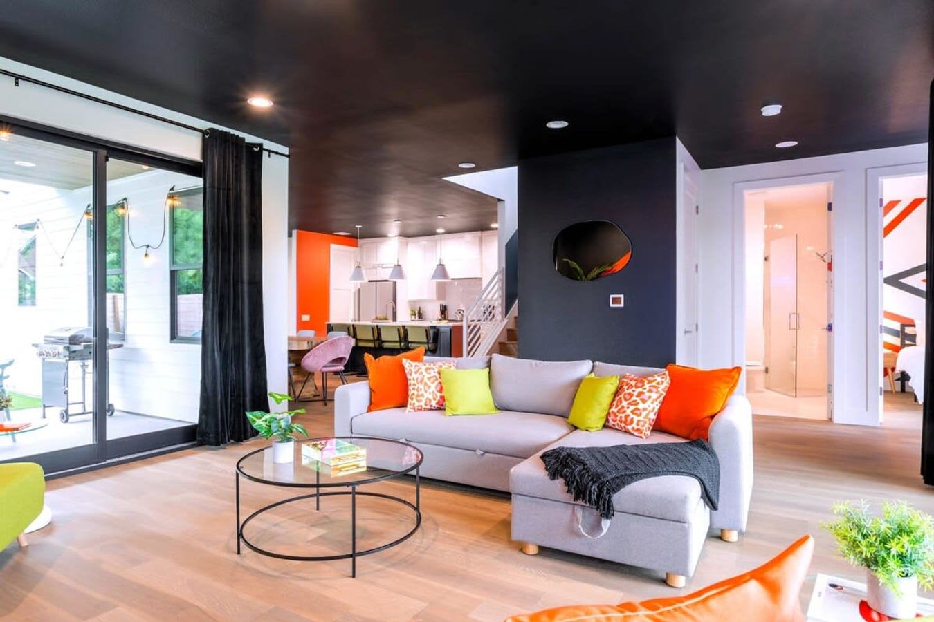 A modern living room features a light gray sectional sofa with colorful orange and lime green pillows, a round glass coffee table, and light wood floors, with a black ceiling and a view into the kitchen and outdoor patio.
