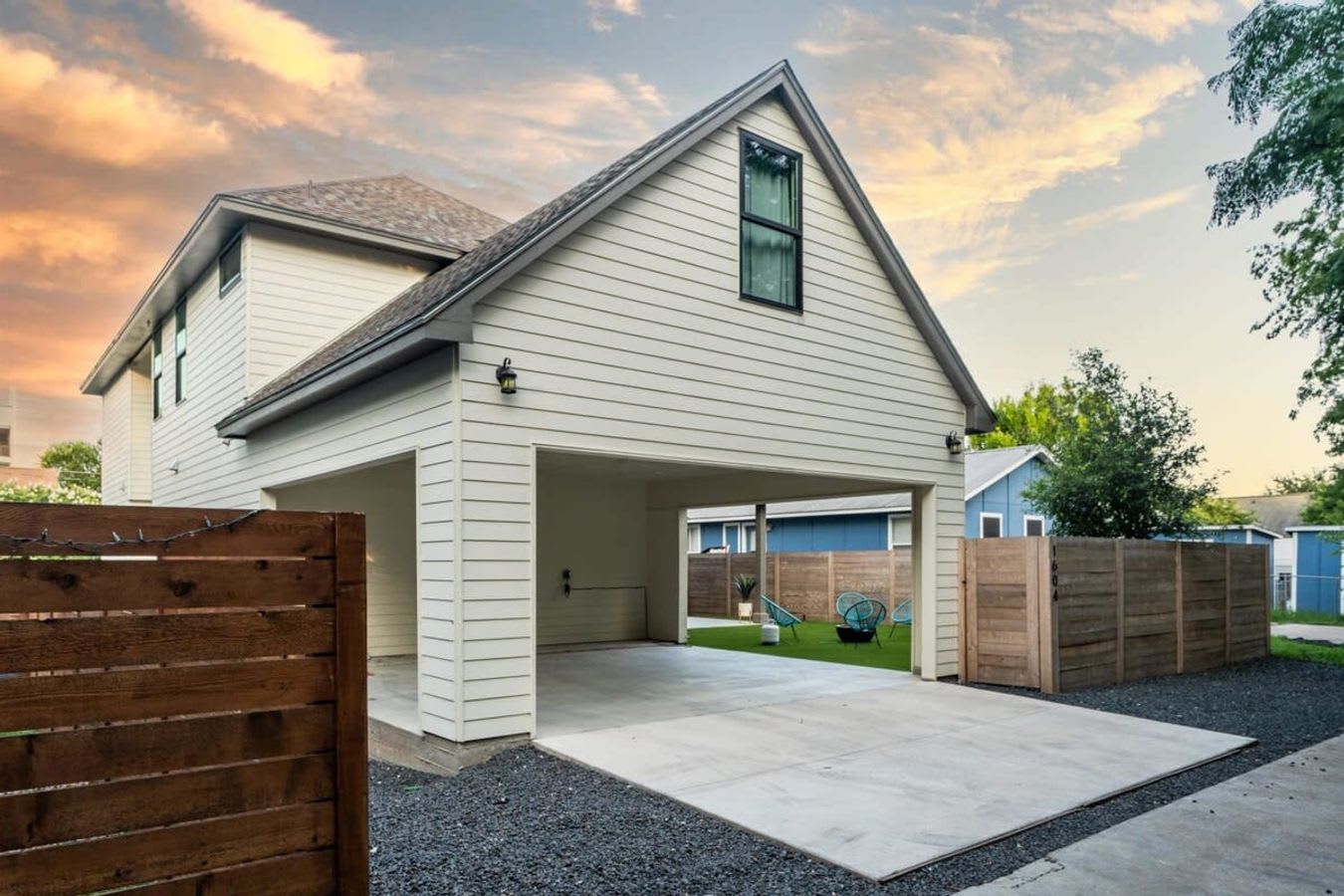 The rear exterior of a modern two-story house with white siding and a gabled roof, featuring a carport or covered parking area. A small fenced yard with artificial turf and outdoor seating is visible to the right.