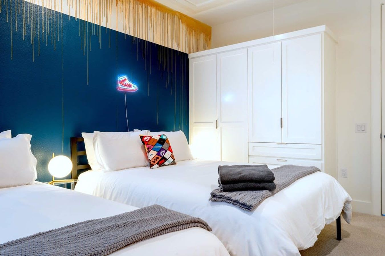 A view of a bedroom with two white beds, one with a gray throw and colorful pillow, against a dark blue wall with gold drip-effect paint and a neon sneaker art piece. A large white built-in wardrobe is visible on the right.