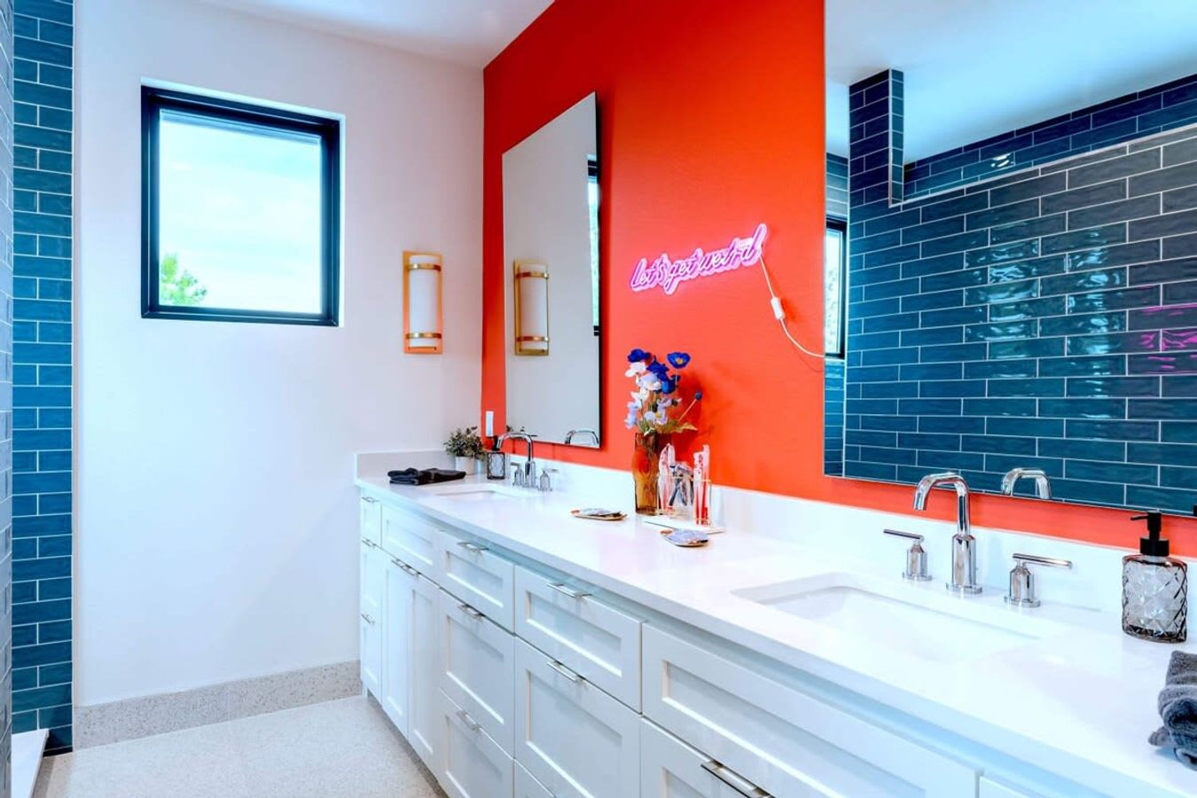 A modern bathroom with a long white double vanity featuring two sinks and chrome faucets. The wall behind the vanity is a vibrant orange with a pink neon sign, contrasting with the white walls and blue subway tile shower visible in the reflection.