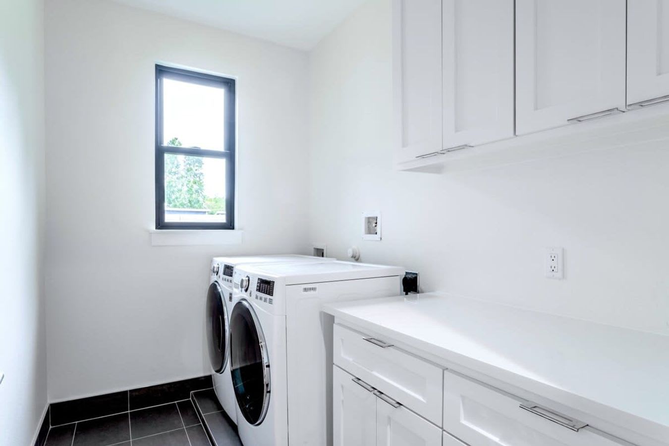 A bright and clean laundry room with white walls, white upper and lower cabinets, and a long white countertop. A front-loading washer and dryer are visible beneath a window, and the floor is tiled in dark grey.