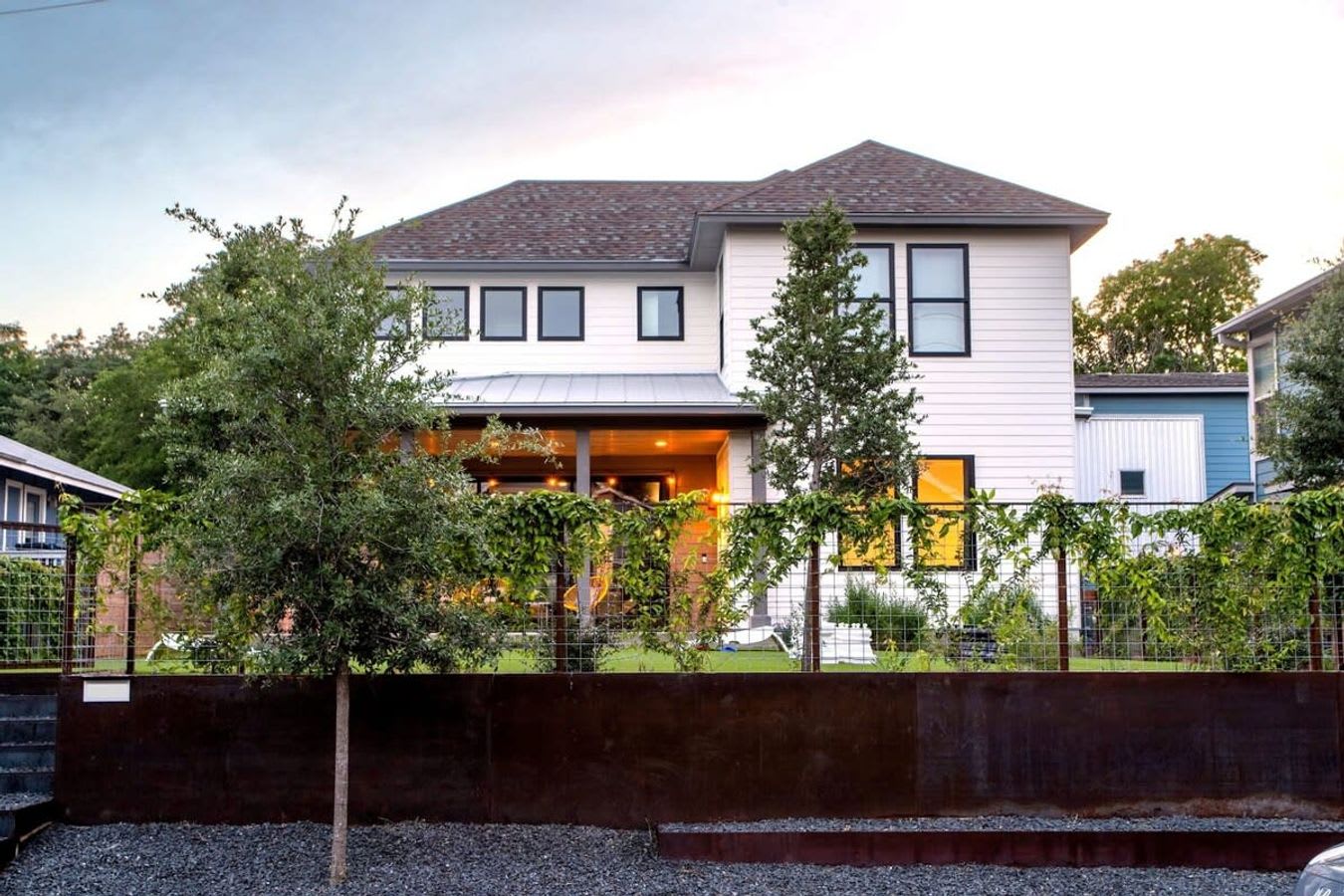 The front exterior of a modern two-story house with white siding and a dark roof, featuring a covered porch and multiple windows. The foreground includes a landscaped area with a metal fence and lush green vines.