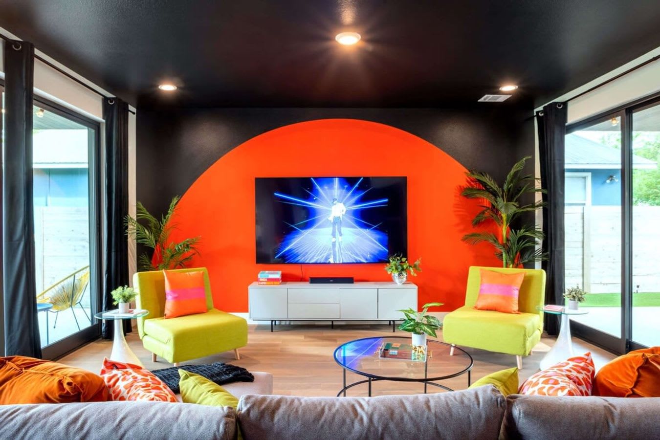 A vibrant living room features a large TV mounted on an orange accent wall with a semi-circular design, flanked by two lime green armchairs and tropical plants, with a gray sectional sofa and black ceiling.