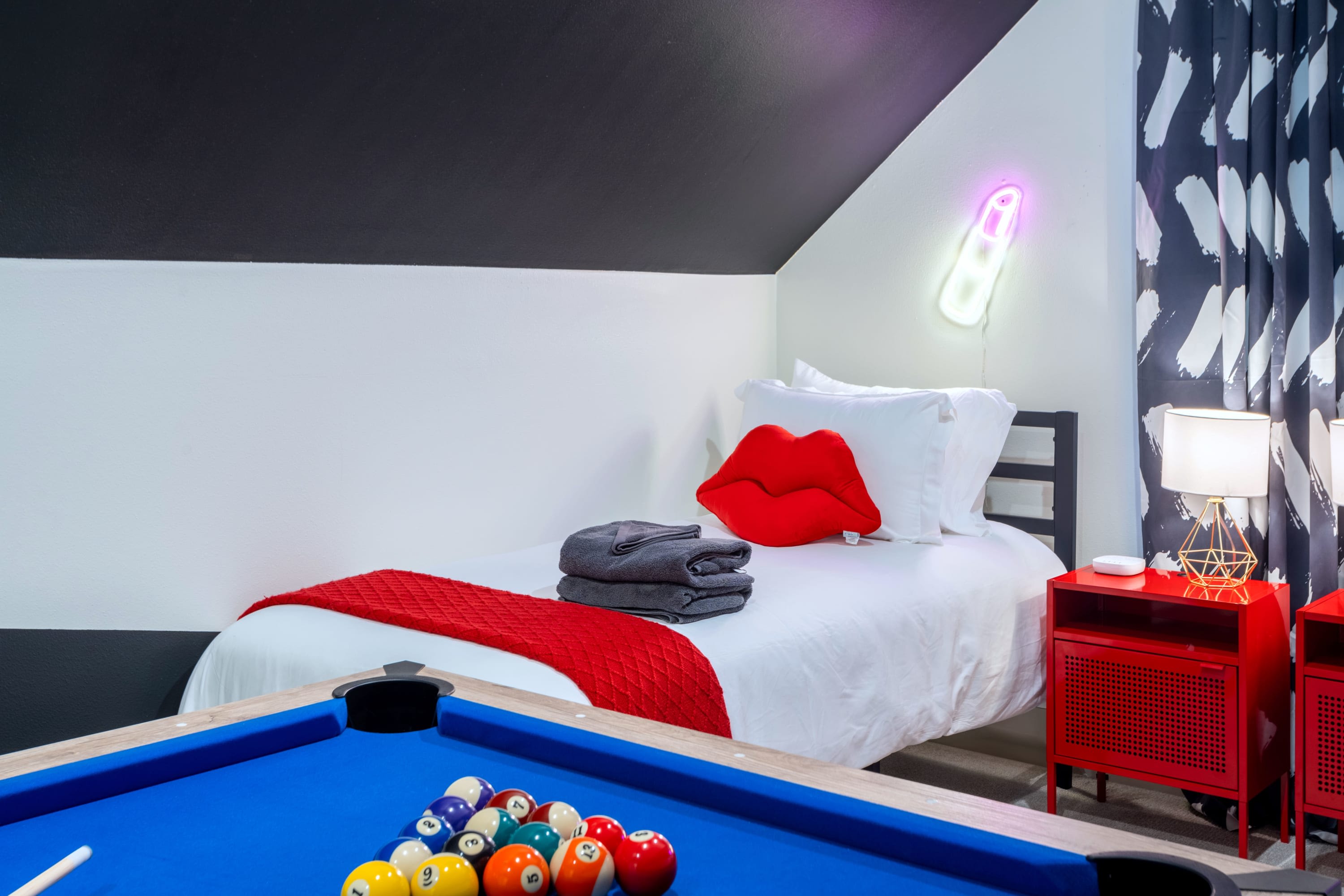 A playful room featuring a single bed with white linens, a red throw, and a red lip-shaped pillow, set against a white wall with bold black diagonal stripes. A blue pool table with colorful balls occupies the foreground, and a pink neon lipstick sign illuminates the wall.