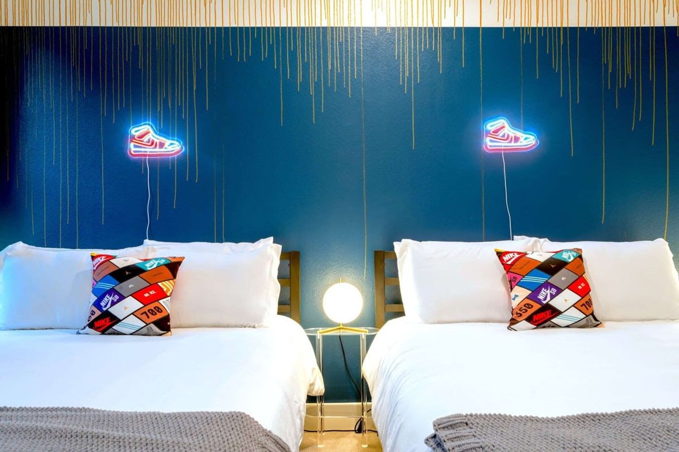 A close-up view of two white beds with colorful patterned pillows and gray throws, against a dark blue wall with gold drip-effect paint. Two glowing neon sneaker art pieces are prominently displayed above the beds.