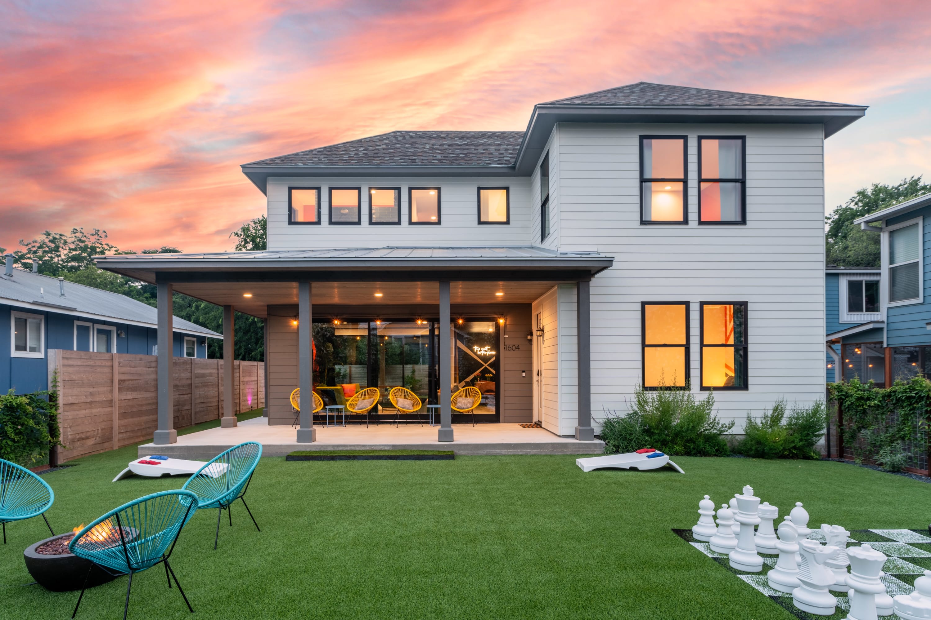The modern white house with dark-framed windows and a covered patio overlooks a lush artificial turf backyard featuring a giant chess set, cornhole boards, and a fire pit area, all under a dramatic, colorful sunset sky.