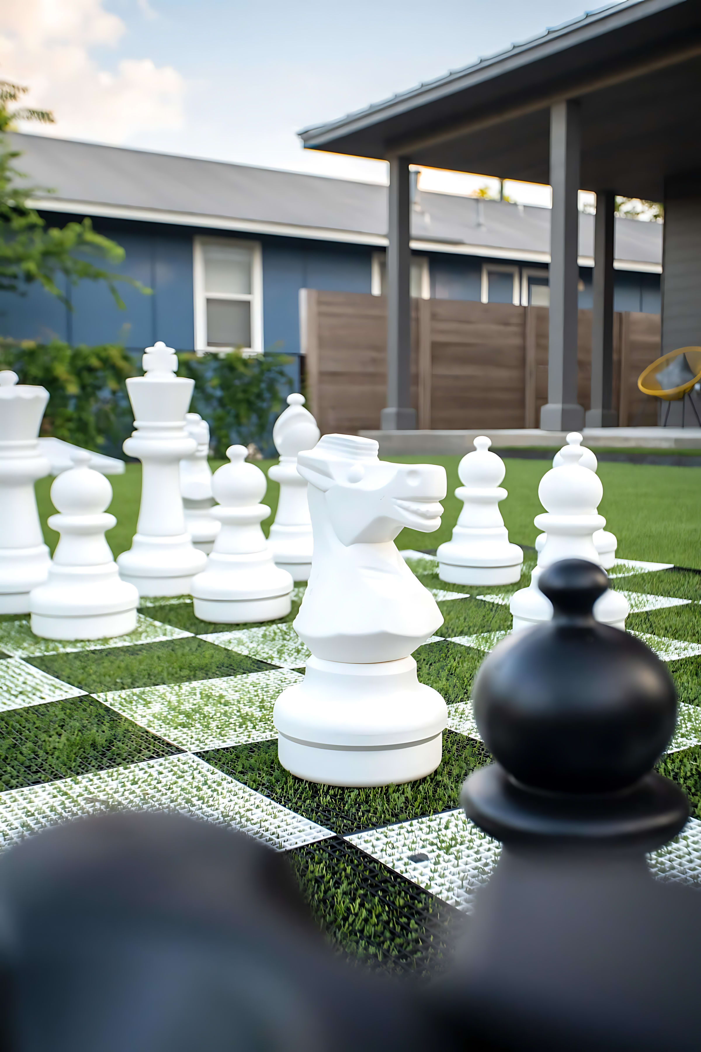 A close-up view of the giant white chess pieces, including a knight, on a green and white checkered mat on artificial turf, with a modern house and covered patio visible in the soft-focus background.