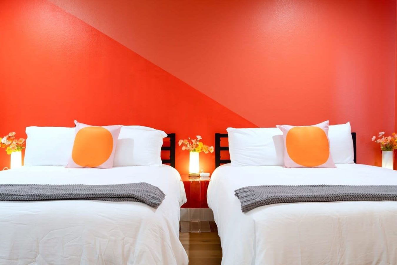 A close-up view of two beds in a vibrant bedroom, each with white linens and an orange circular pillow, flanked by small nightstands with lamps. The striking orange accent wall features a diagonal lighter orange stripe.