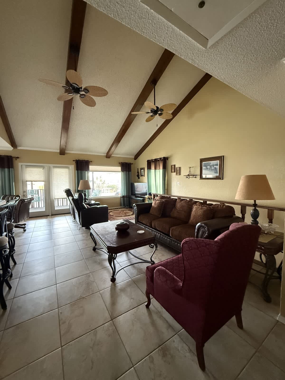 A spacious living room with a high vaulted ceiling, exposed wooden beams, and two ceiling fans. It features a brown sofa, a red armchair, and a wooden coffee table, with large windows overlooking the exterior.