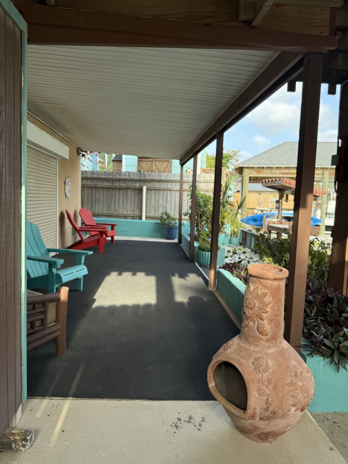 A ground-level covered patio with a concrete floor and dark outdoor carpet, furnished with colorful Adirondack chairs and an outdoor chiminea, leading to a garden area.