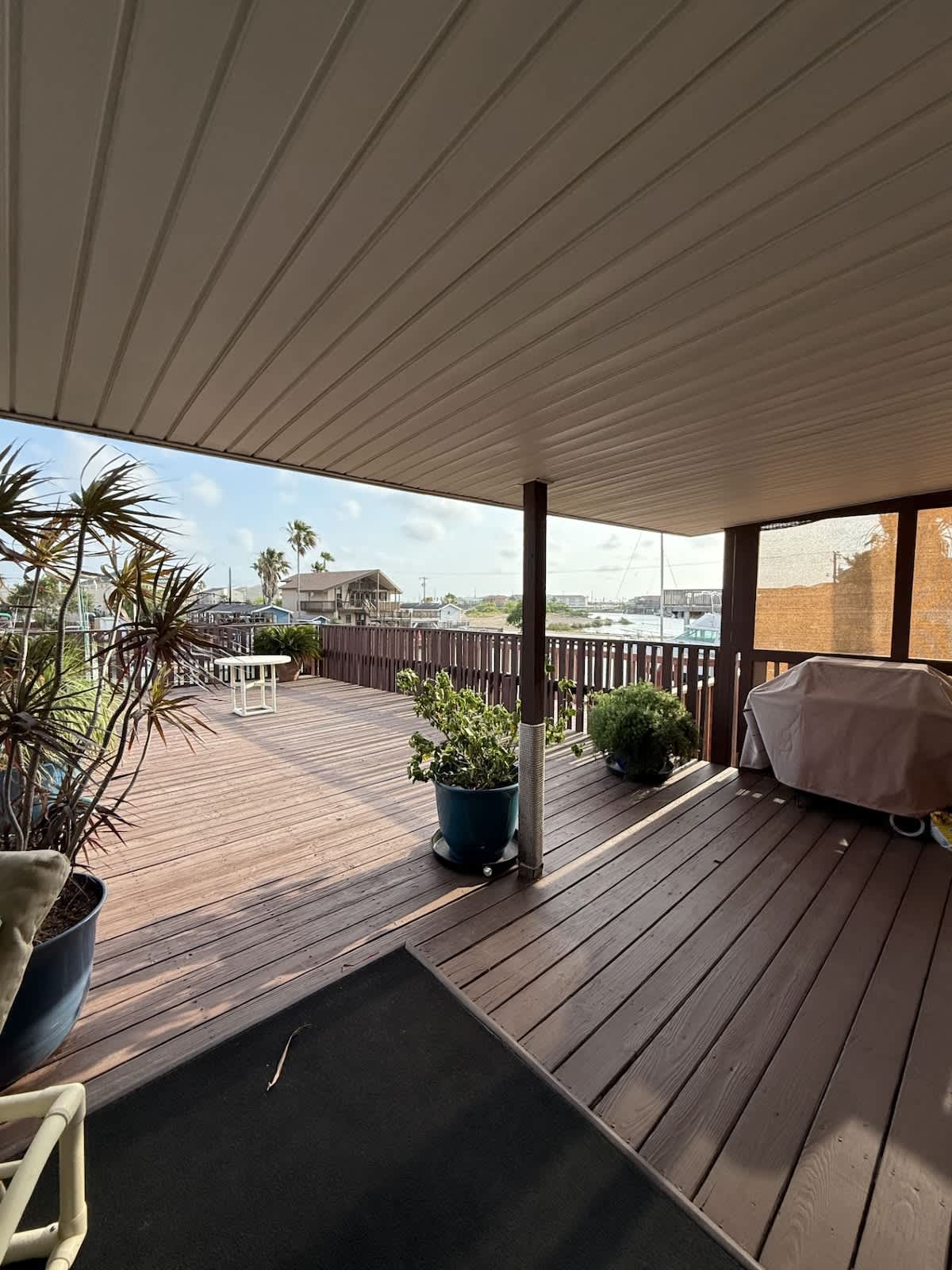 A spacious wooden deck overlooking a canal with boats, featuring several large potted plants, a small white table, and a sturdy wooden railing under a partly cloudy sky.