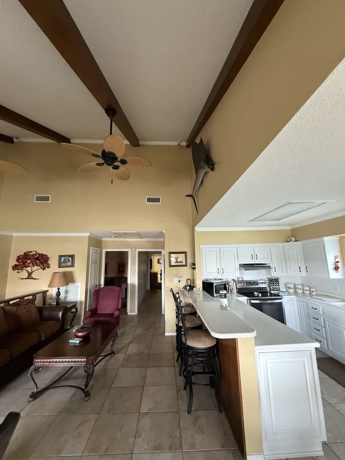 An expansive open-concept living room and kitchen area with a high vaulted ceiling and exposed wooden beams. The living space includes a brown sofa and a red armchair, while the kitchen features white cabinets and a breakfast bar.