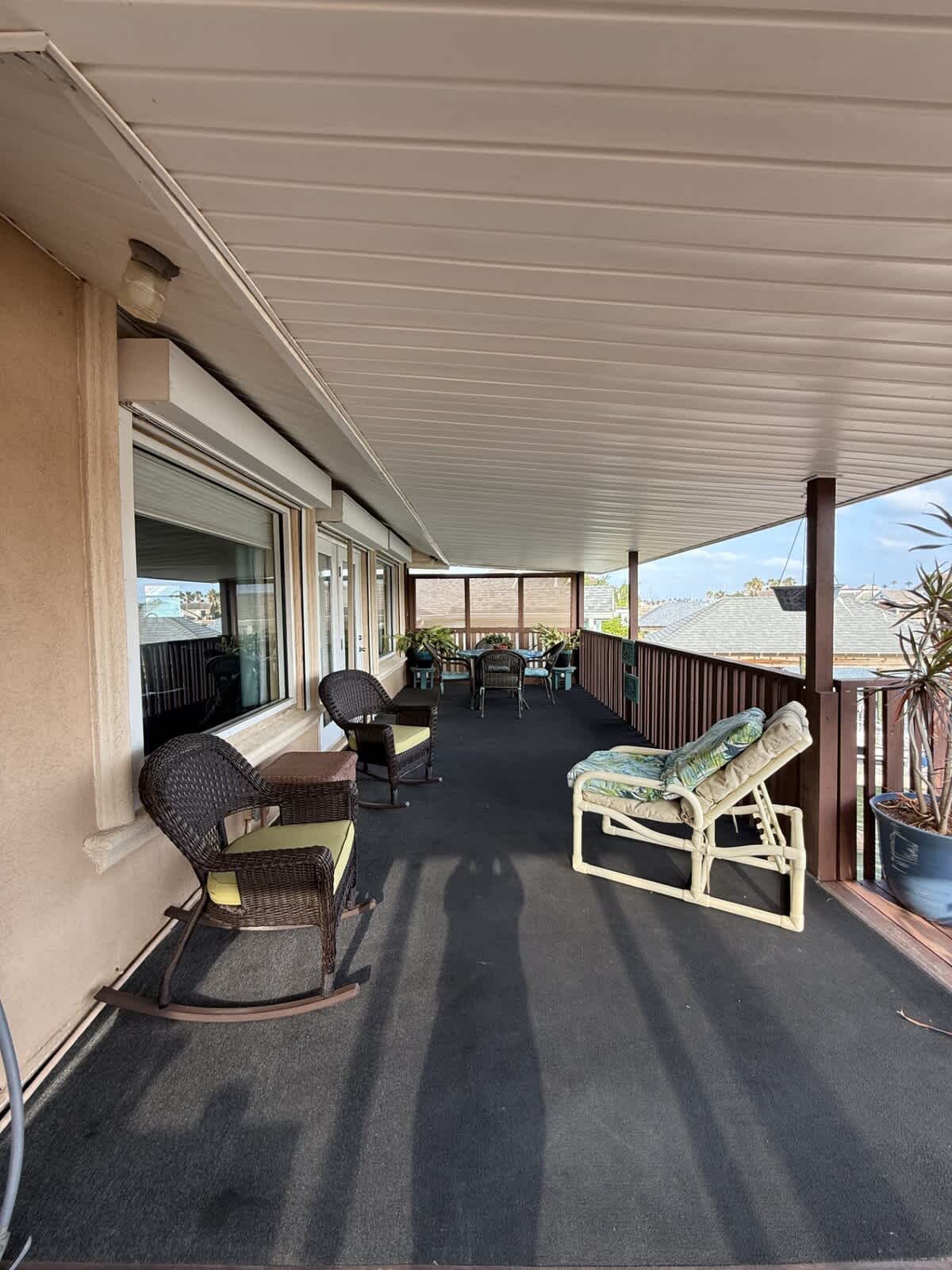 A long, covered outdoor patio with dark carpet, furnished with wicker rocking chairs, side tables, and a lounge chair, offering a view of the water and a distant grill area.