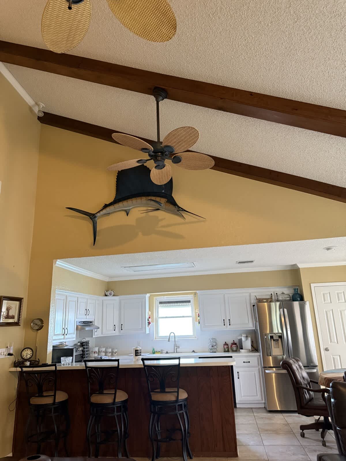 A view of the open-concept living area and kitchen, featuring a high vaulted ceiling with exposed wooden beams and a large mounted fish decoration. The kitchen area includes white cabinets and a breakfast bar.