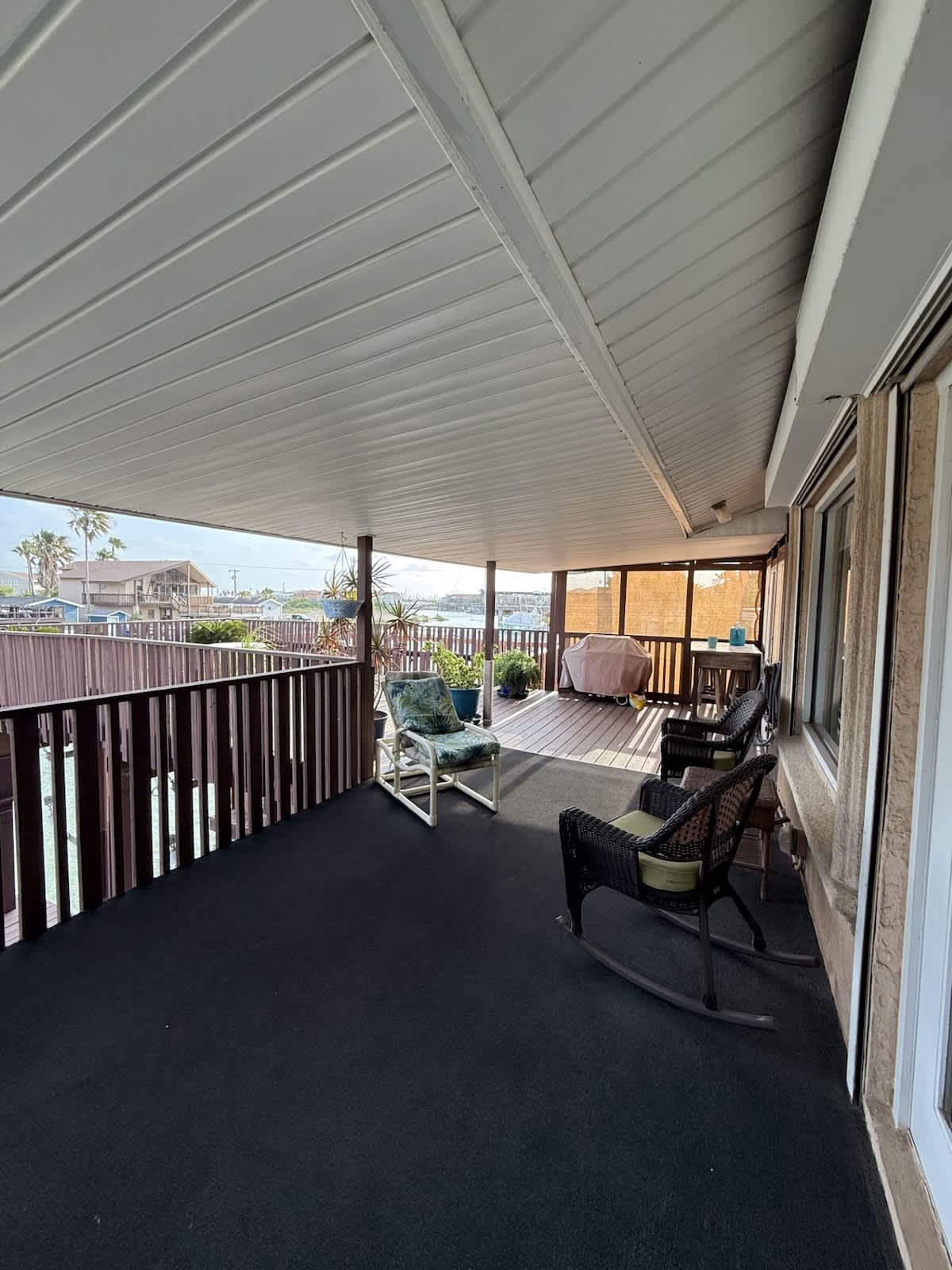 A wide, covered patio with dark outdoor carpeting, featuring wicker rocking chairs and a lounge chair, providing an open view of the water and the adjacent wooden deck.