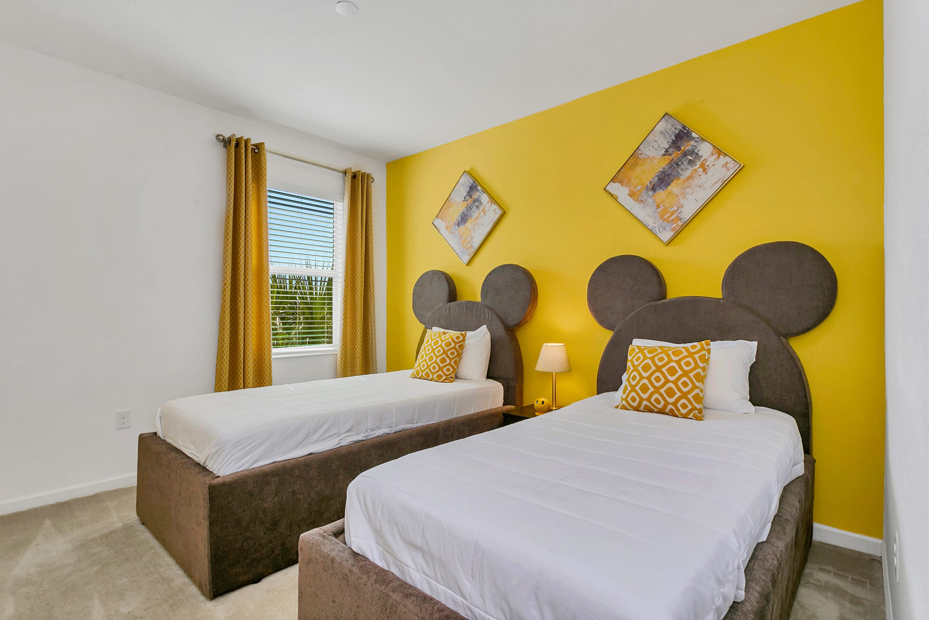 A vibrant children's bedroom featuring two twin beds with unique Mickey Mouse ear-shaped headboards, set against a bright yellow accent wall adorned with two abstract art pieces. A window with blinds and golden curtains is visible on the left.