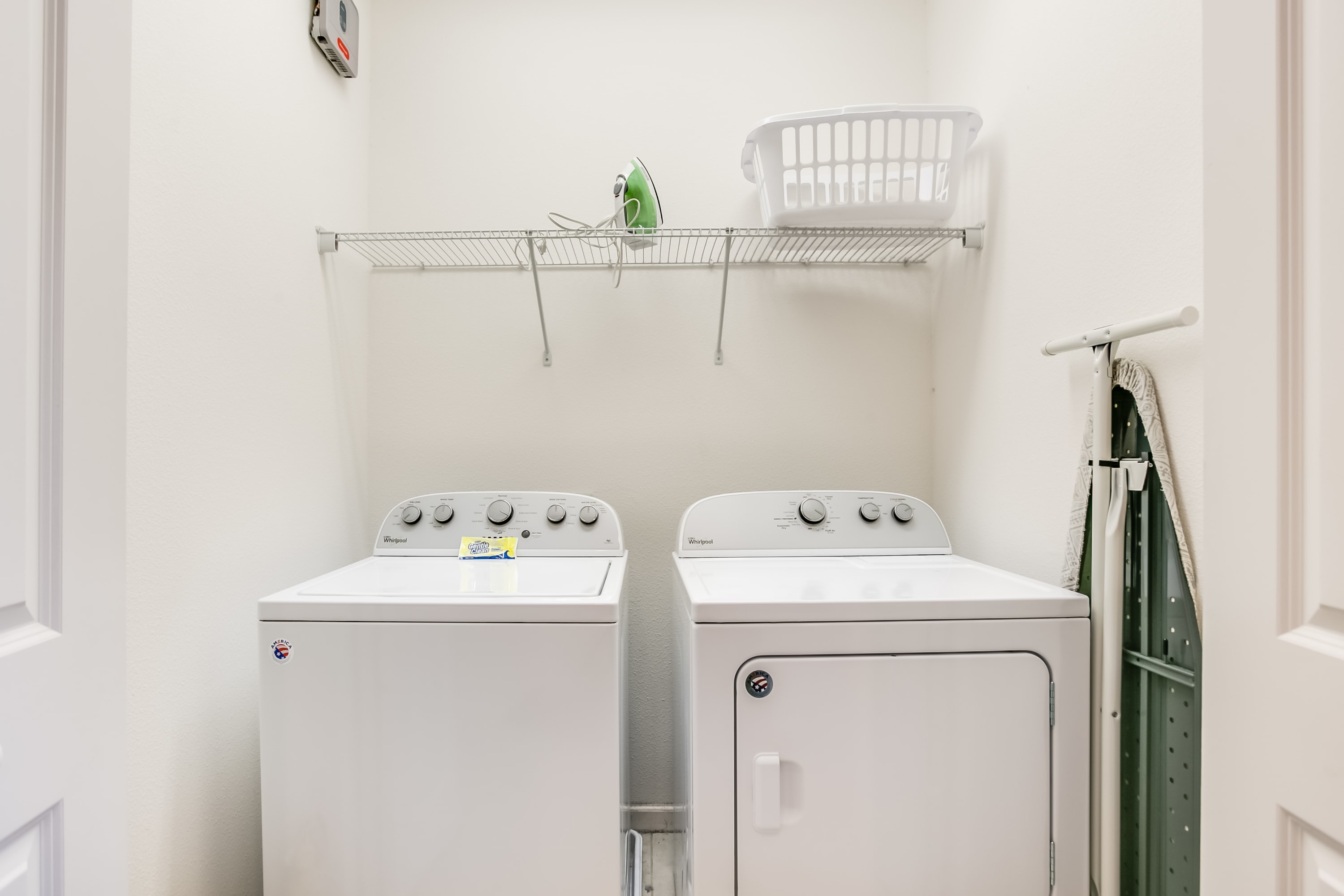 A functional laundry room equipped with a white Whirlpool top-load washing machine and a matching front-load dryer, positioned beneath a wire shelf holding an iron and a laundry basket.
