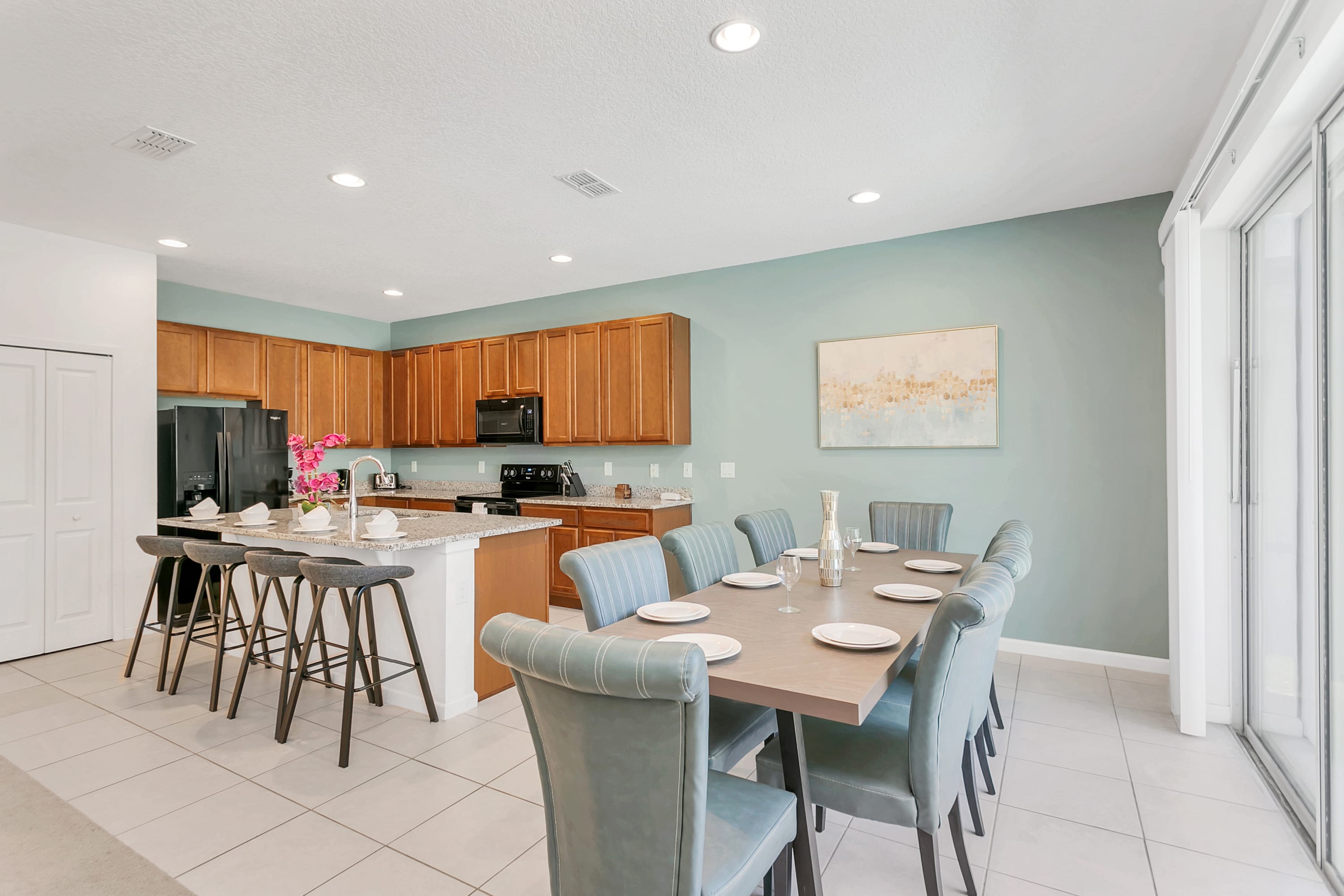 A spacious open-concept kitchen with light wood cabinetry, granite countertops, and black appliances, including a large island with bar seating. Adjacent is a dining area with a long wooden table and light blue upholstered chairs, all on white tile flooring.