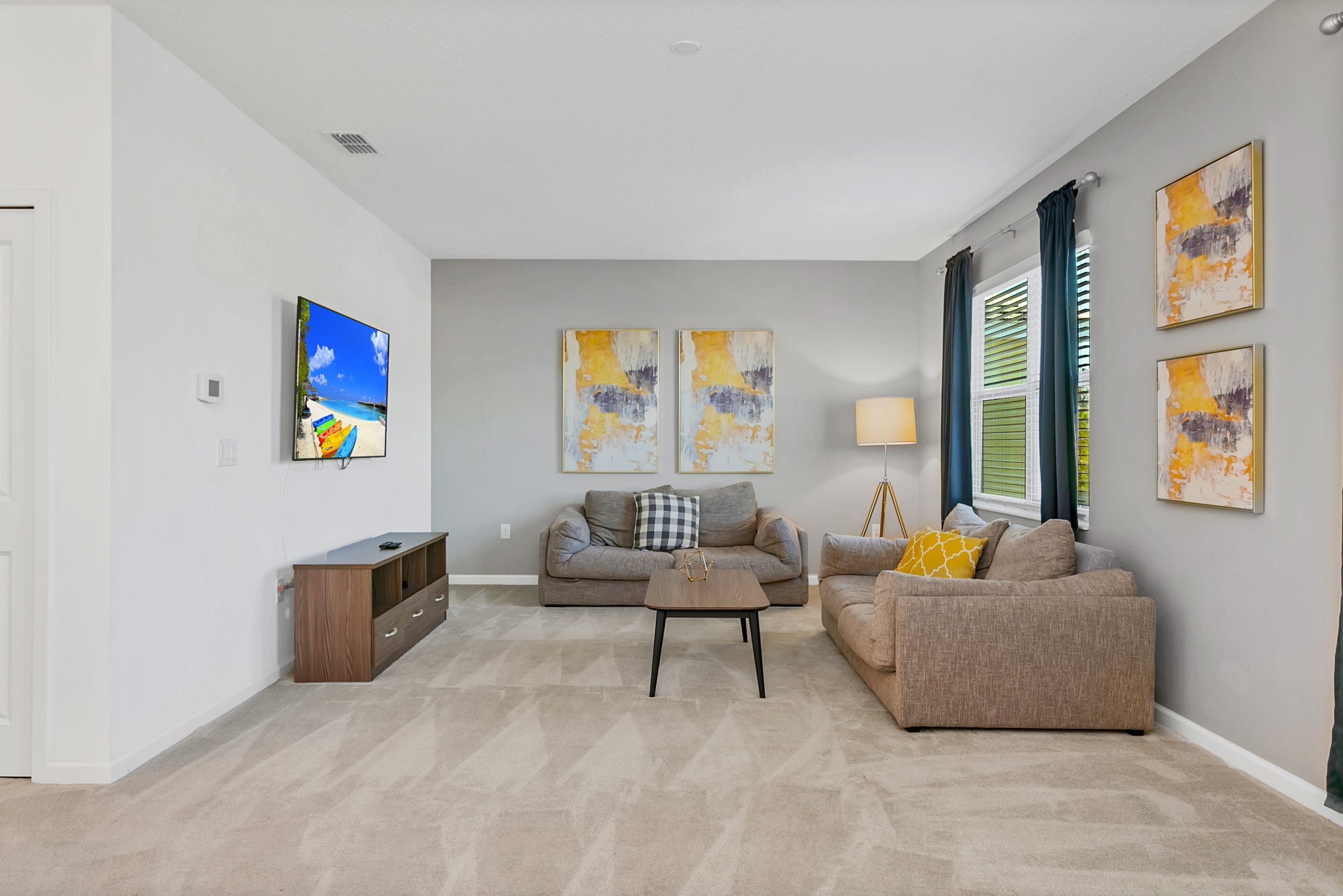 A bright living room with light gray walls and carpet, featuring two neutral-toned sofas, a dark wood coffee table, and a wall-mounted TV. Two abstract yellow and gray art pieces adorn the main wall, with a window on the right.