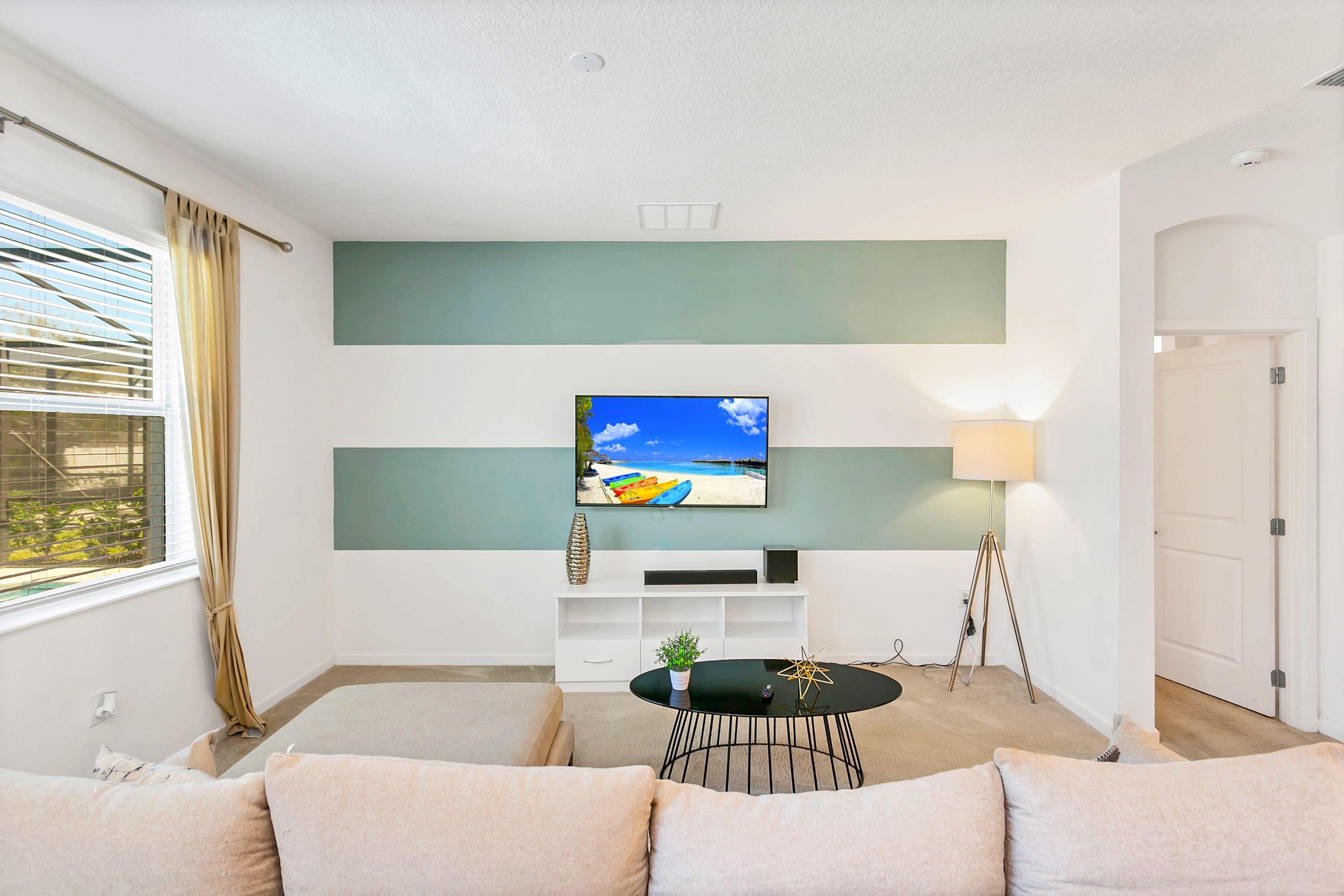A bright living room with a large wall-mounted TV displaying a beach scene, set against a white wall with two horizontal light blue stripes. A beige sectional sofa is partially visible, along with a black coffee table and a floor lamp.