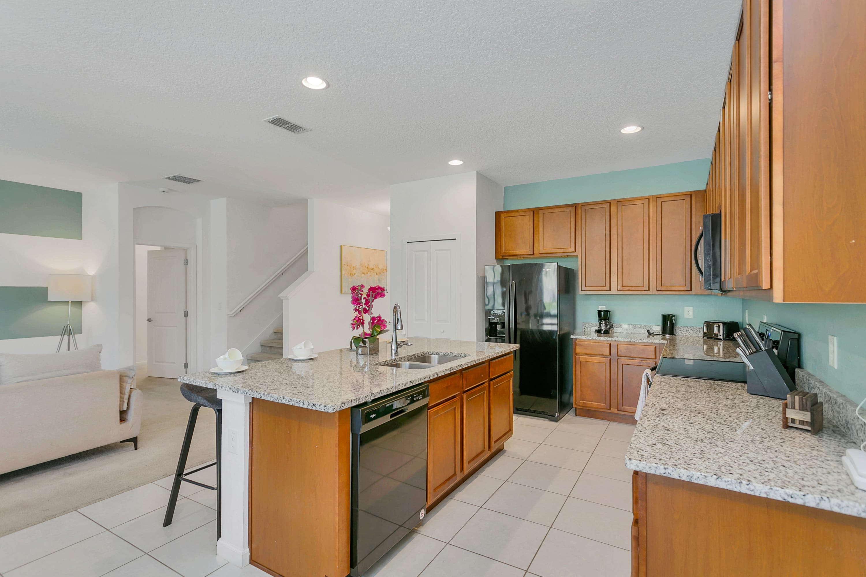 A well-equipped kitchen featuring light wood cabinets, speckled granite countertops, and black appliances including a refrigerator, microwave, and dishwasher. A kitchen island with a sink and bar stools is prominent, and the floor is white tile.