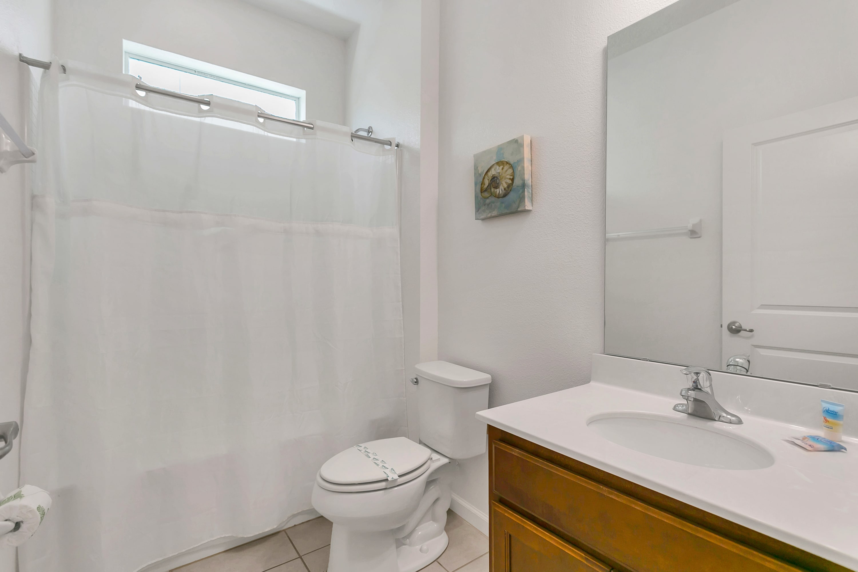 A clean, white bathroom featuring a toilet with a closed lid, a shower/tub combo enclosed by a white curtain, and a wooden vanity with a white countertop and sink, all under a small window.