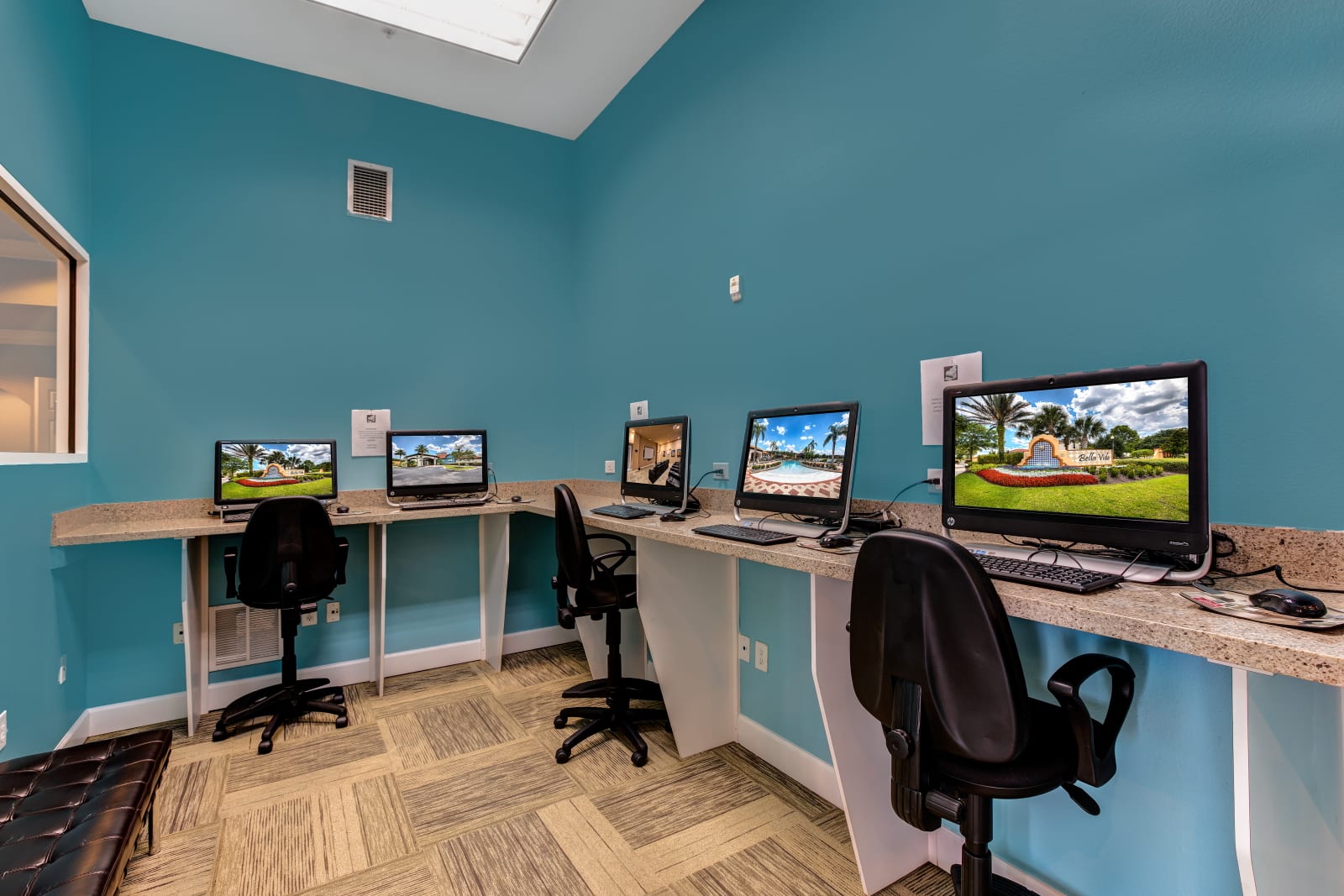 A communal business center with teal walls, offering multiple computer workstations with desktop monitors and chairs, set along a long countertop, providing a dedicated space for work or internet access.