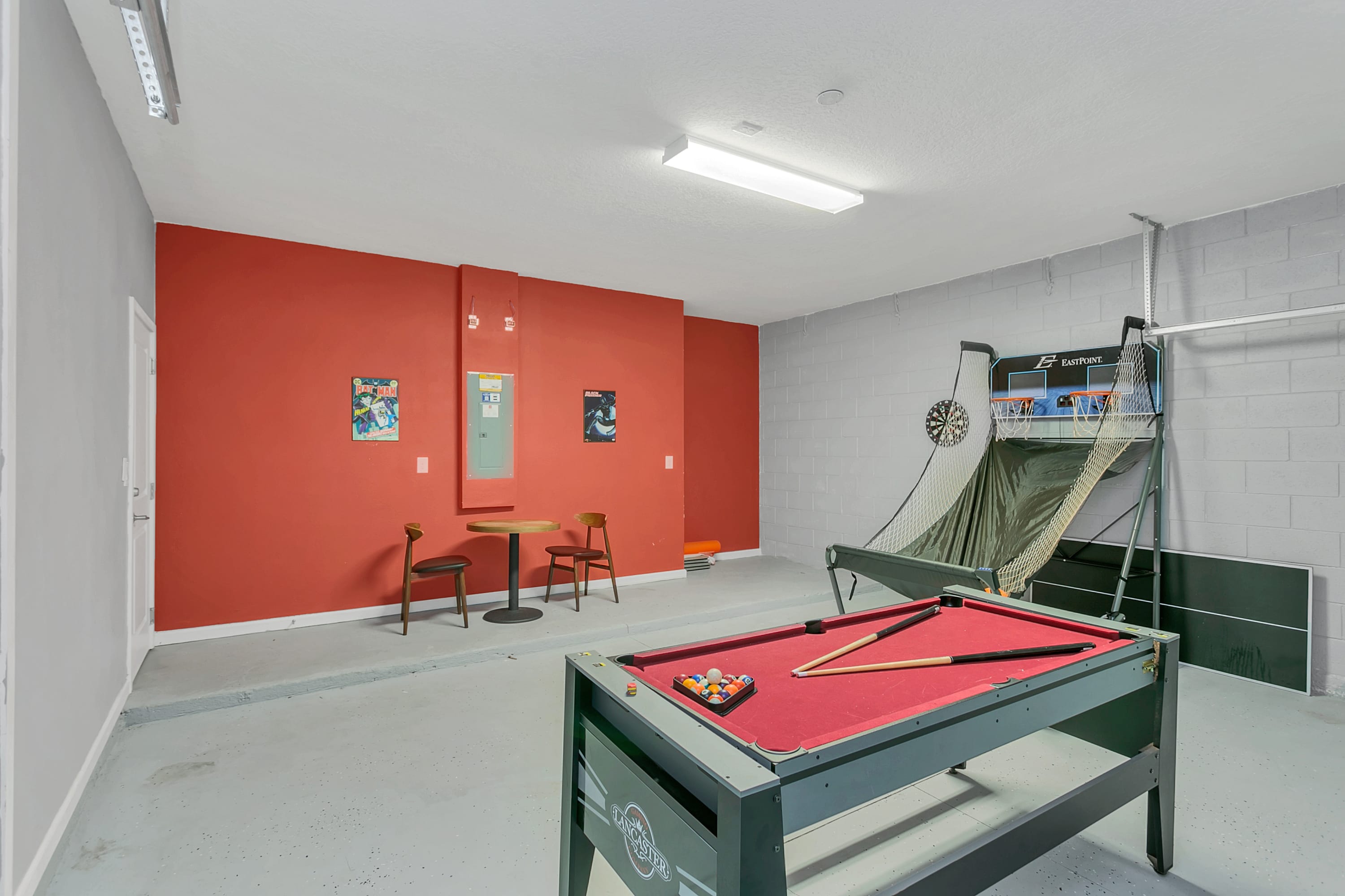 A vibrant game room with a red accent wall, featuring a red-felt pool table in the foreground, a basketball arcade game, and a dartboard. A small round table with two chairs is placed against the red wall, and the floor is a grey speckled concrete.