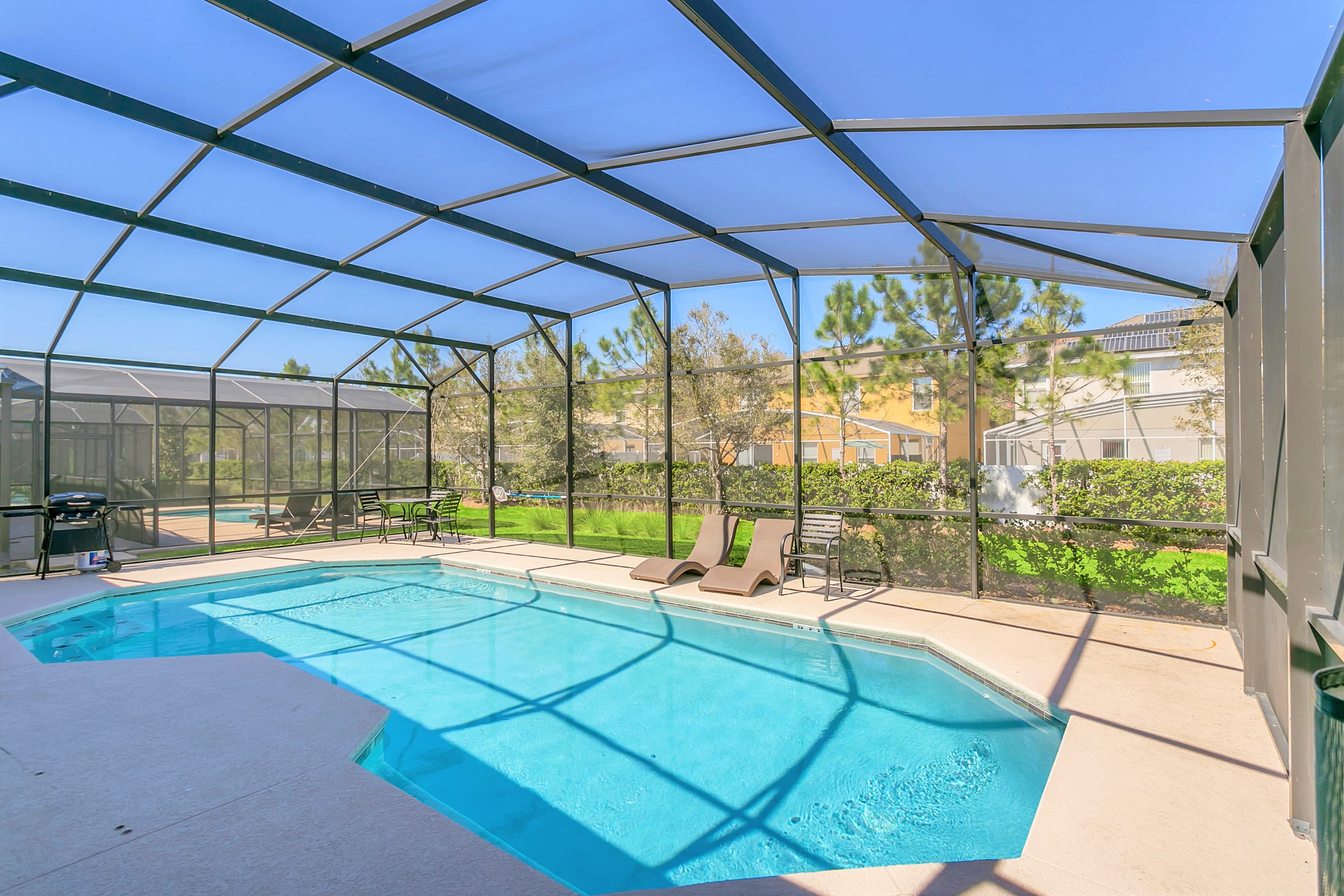 A bright blue swimming pool enclosed within a large screen cage, featuring two brown lounge chairs on the concrete deck and a small dining set with a grill in the background. The pool is surrounded by a concrete patio and overlooks a grassy yard and neighboring houses.
