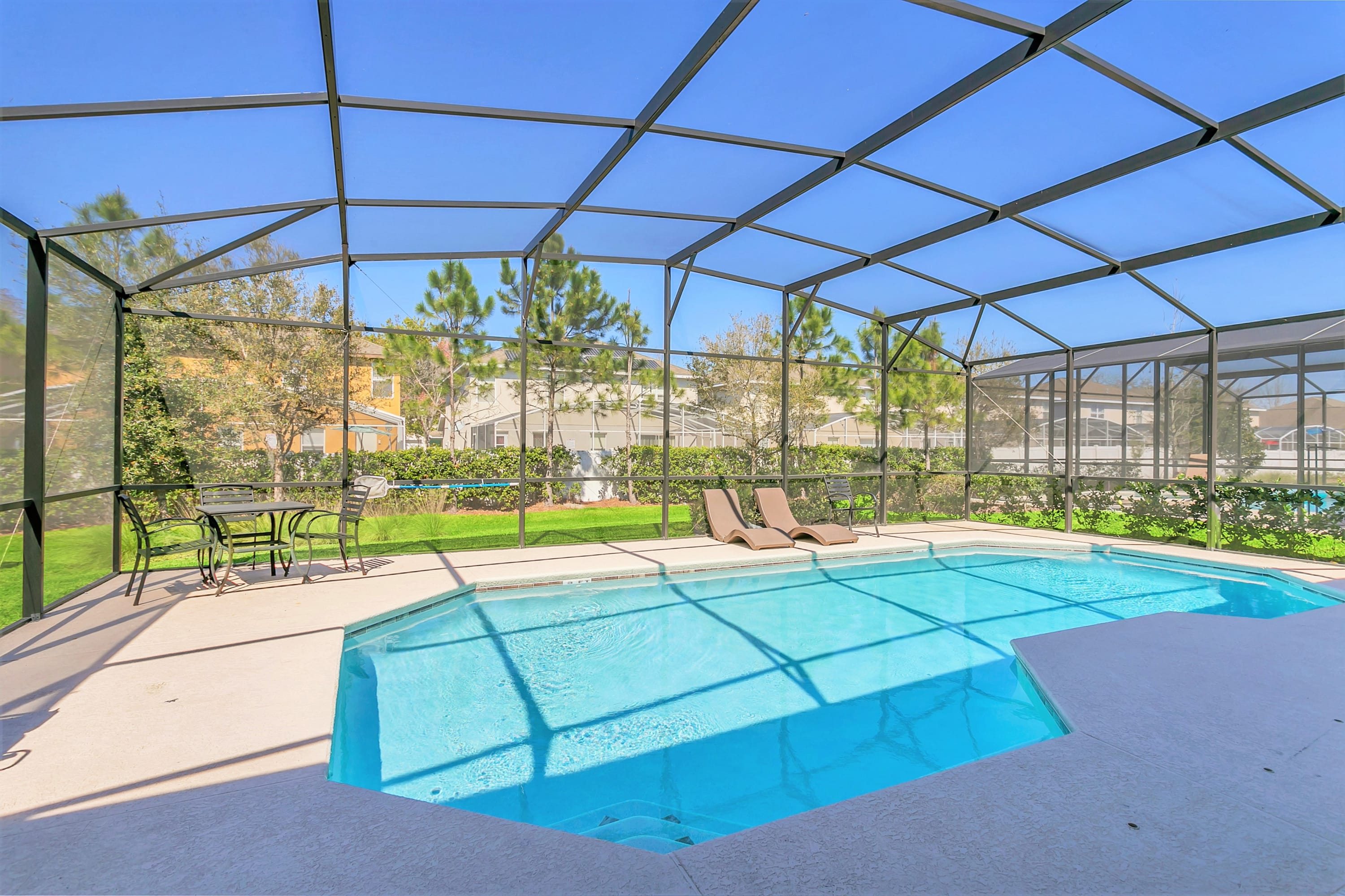 A sparkling blue swimming pool enclosed within a large screen cage, featuring two brown lounge chairs on the concrete deck. The pool area overlooks a well-maintained grassy yard and residential homes in the distance under a clear blue sky.