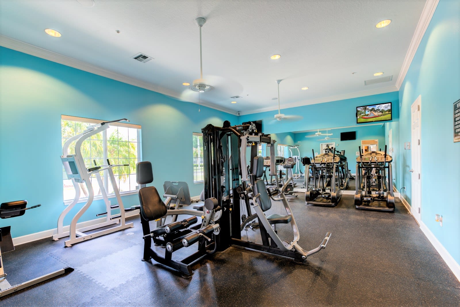A well-equipped fitness center with bright blue walls, featuring various exercise machines including a multi-station weight machine, treadmills, and ellipticals, with large mirrors and ceiling fans.