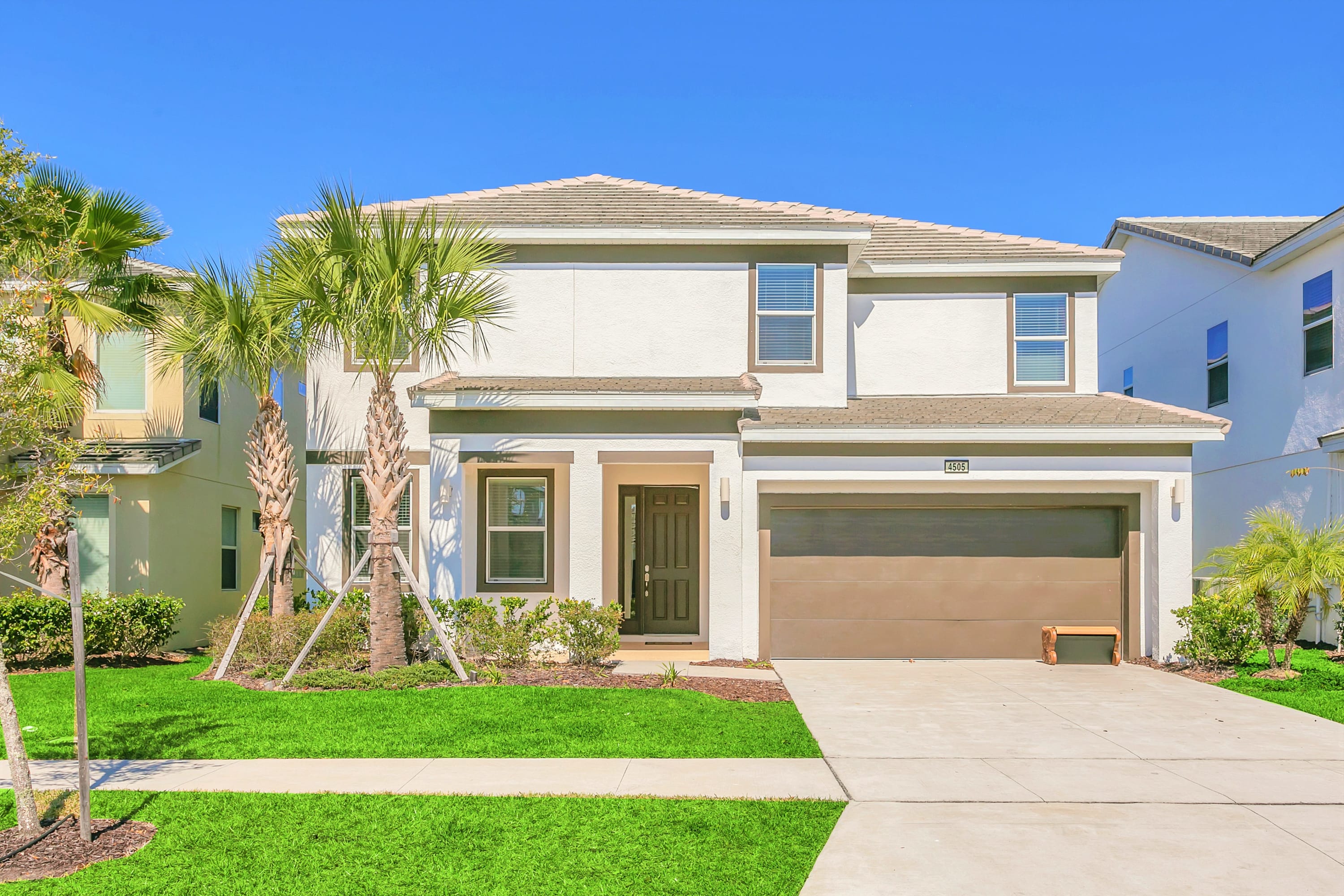 A modern two-story house with a light-colored stucco exterior and a brown garage door, set against a clear blue sky. The front yard features a well-maintained green lawn, palm trees, and a concrete driveway leading to the garage.