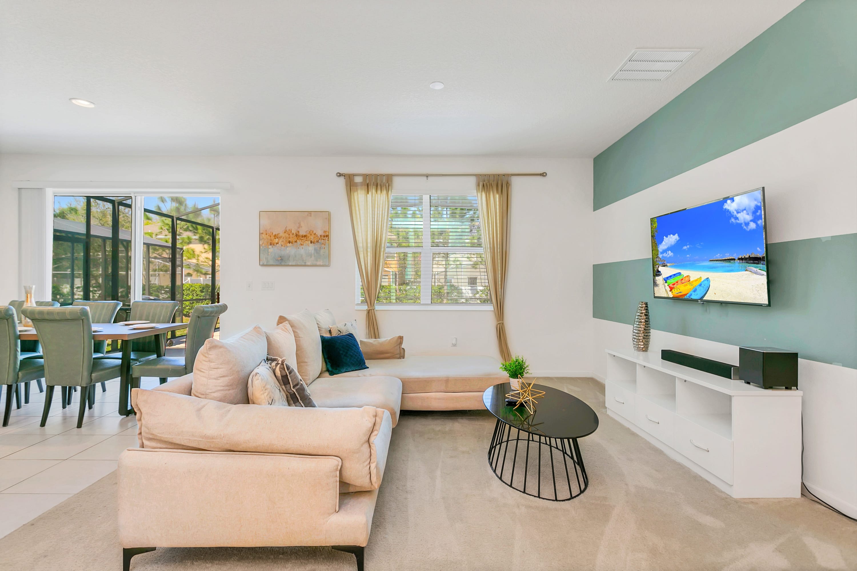 A cozy living room featuring a large beige sectional sofa, a black coffee table, and a wall-mounted TV above a white media console. The wall behind the TV has horizontal stripes in white and light blue, and a window with blinds is visible on the left.